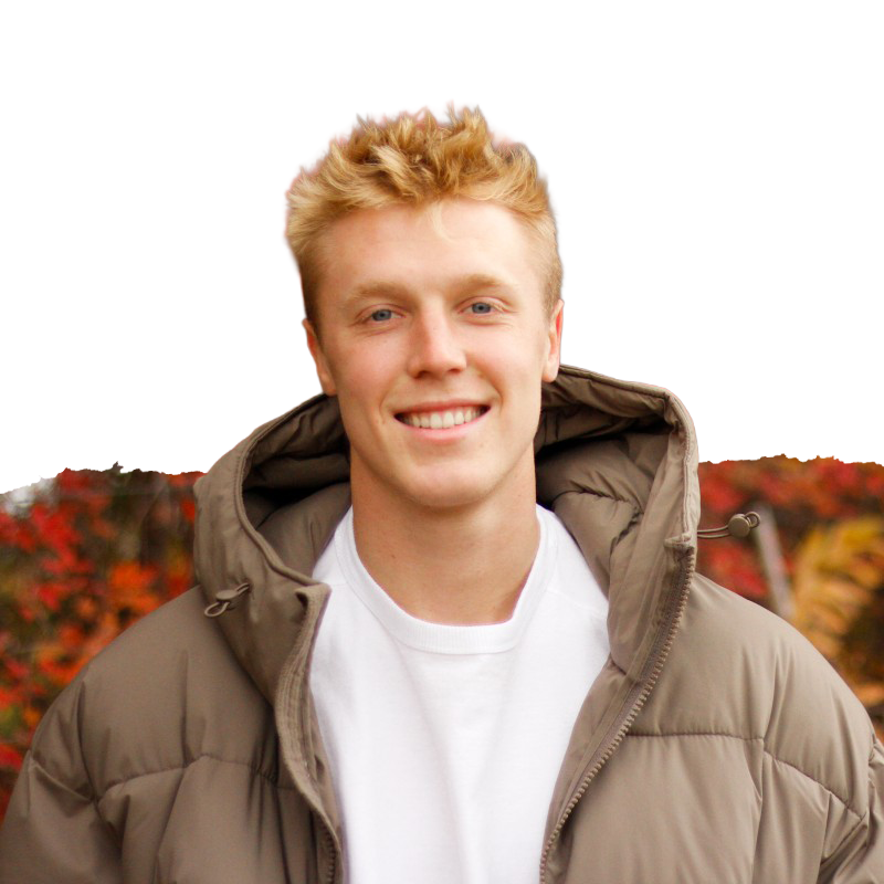 A young man with blonde curly hair smiling outdoors during autumn, wearing a tan puffer jacket and a white t-shirt.