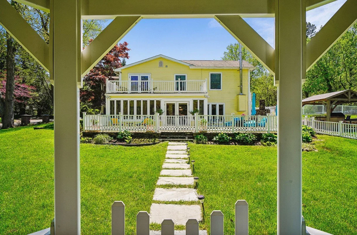 View of a yellow house with multiple decks and a fenced backyard, seen from under a porch with white pillars, on a sunny day with green trees and a well-maintained lawn.