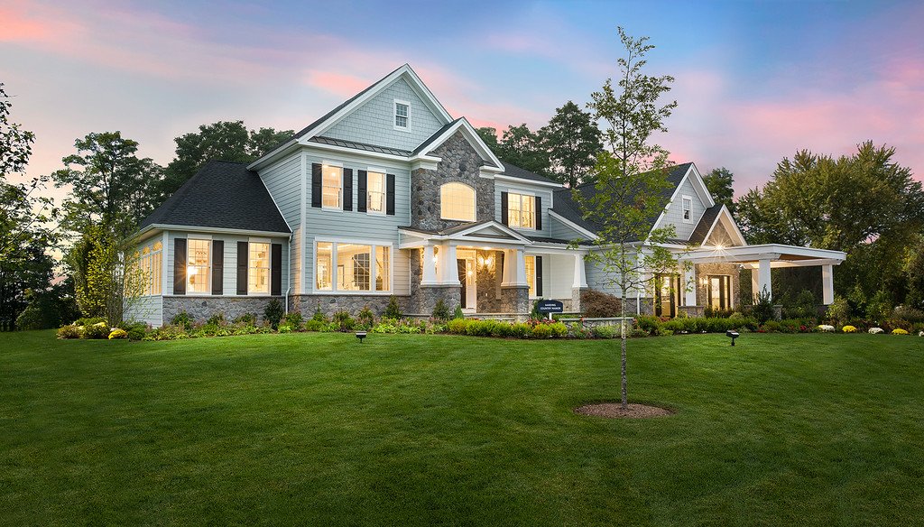 Large, elegant house with white siding and stone accents, surrounded by a well-maintained lawn and trees, during sunset.