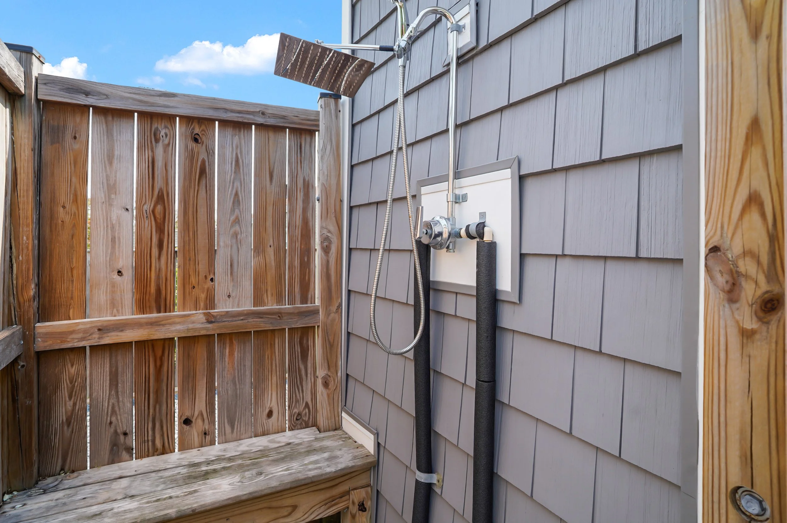 Outdoor shower with metallic showerhead, connected to gray exterior siding, next to a wooden fence under a blue sky with clouds.
