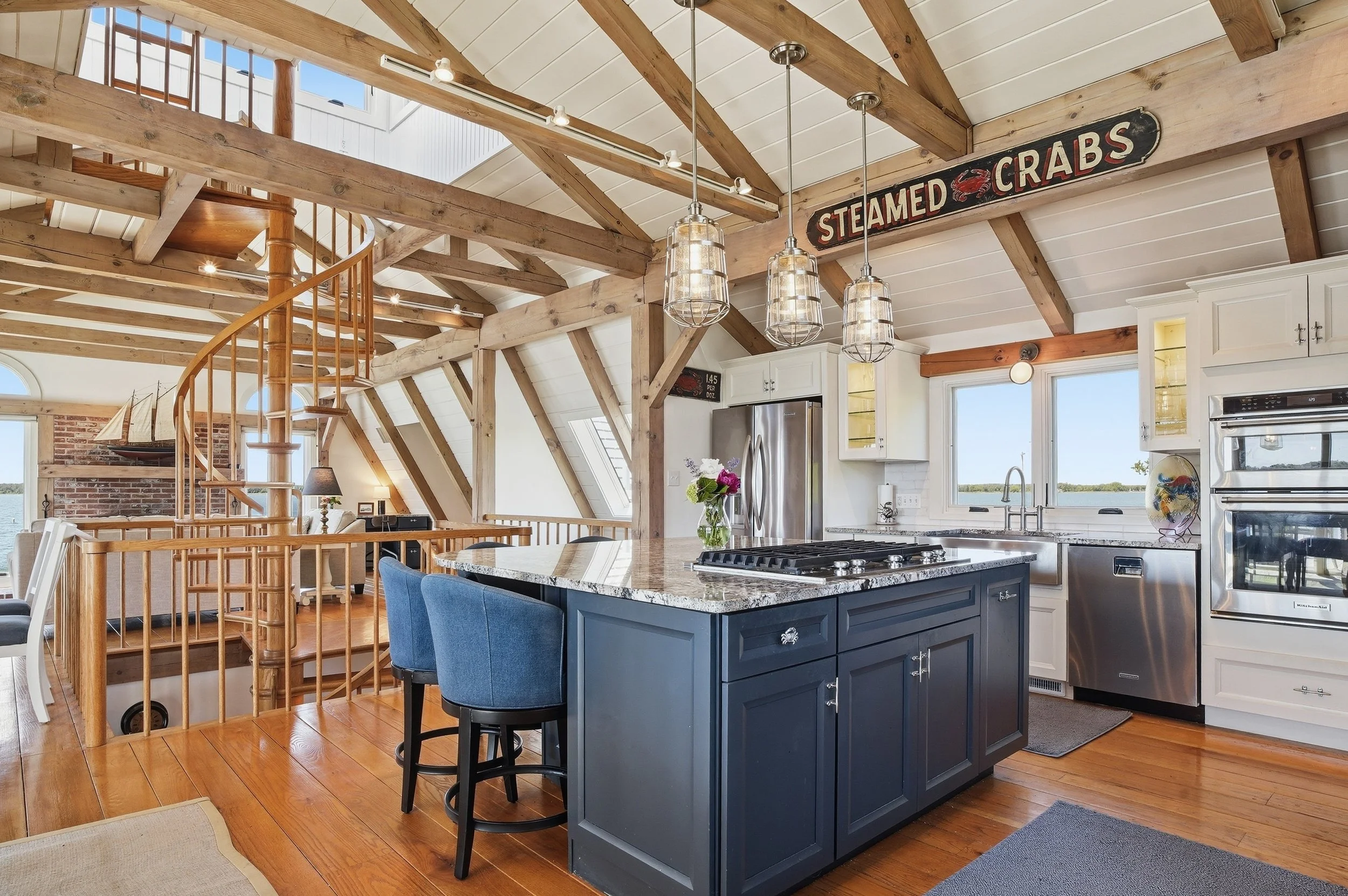 Open-concept kitchen with vaulted ceilings, exposed wood beams, white cabinetry, stainless steel appliances, and a central dark blue island with a granite countertop. There is a spiral staircase in the background leading to an upper level, and a sign