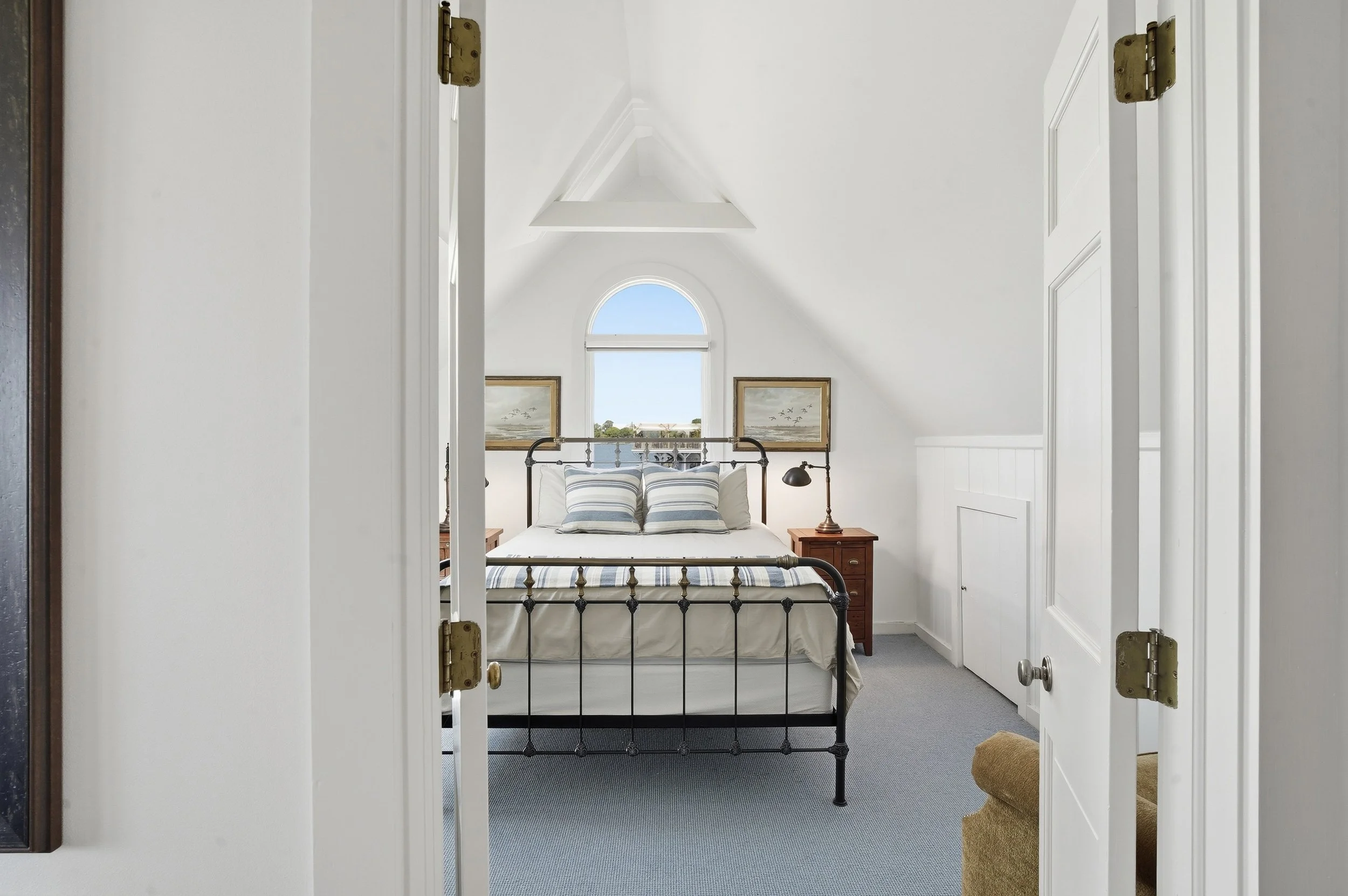 View of a cozy bedroom through a doorway, featuring a black iron bed frame with a white bed and striped pillows, two wooden nightstands with lamps, framed artwork, and a large arched window with a view of the sky and trees outside.