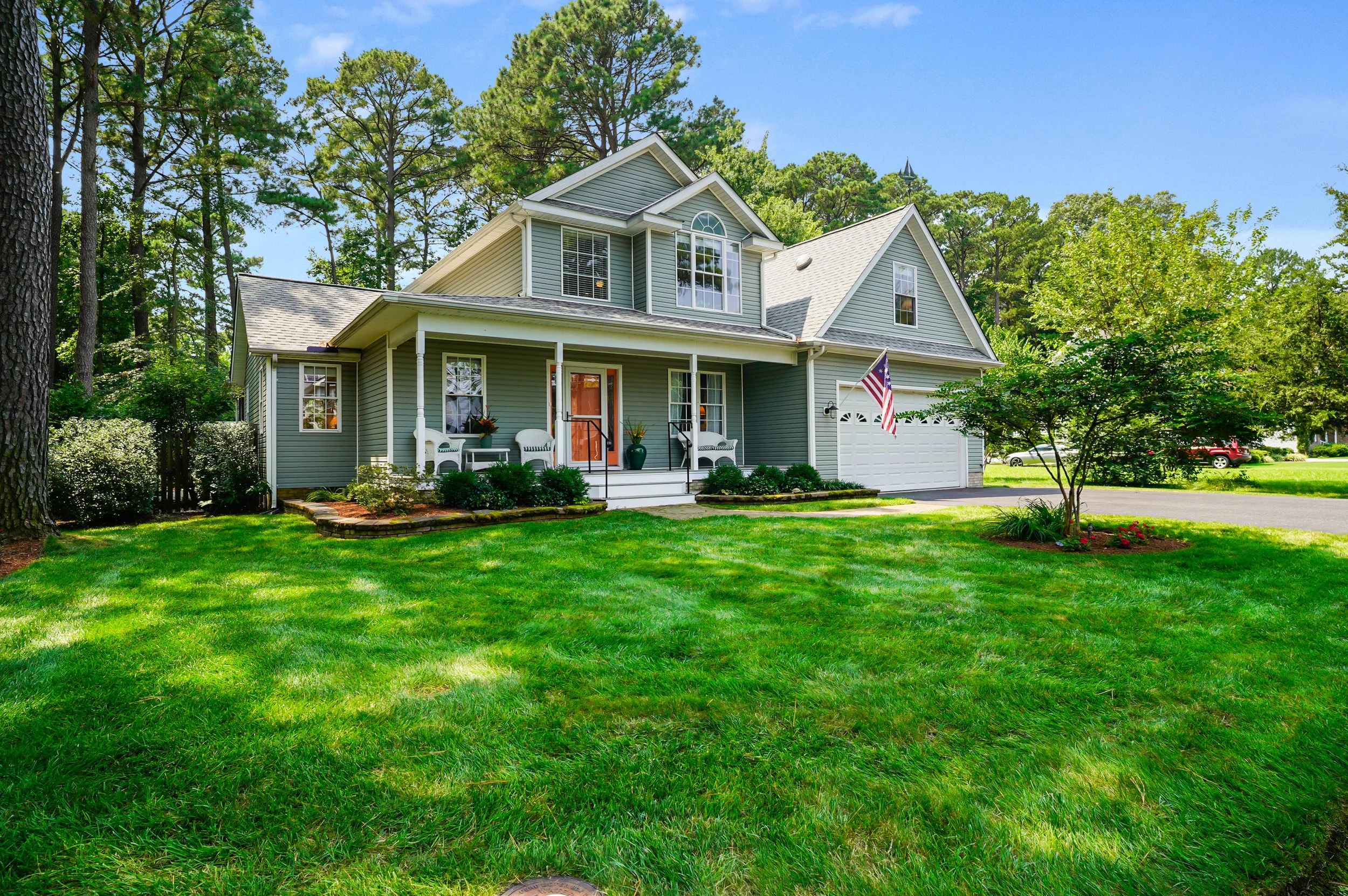 A two-story green house with a front porch, white railing, benches, and potted plants. The house has multiple large windows and an American flag hanging outside. The lawn is green with small trees and shrubbery, surrounded by tall pine trees under a 