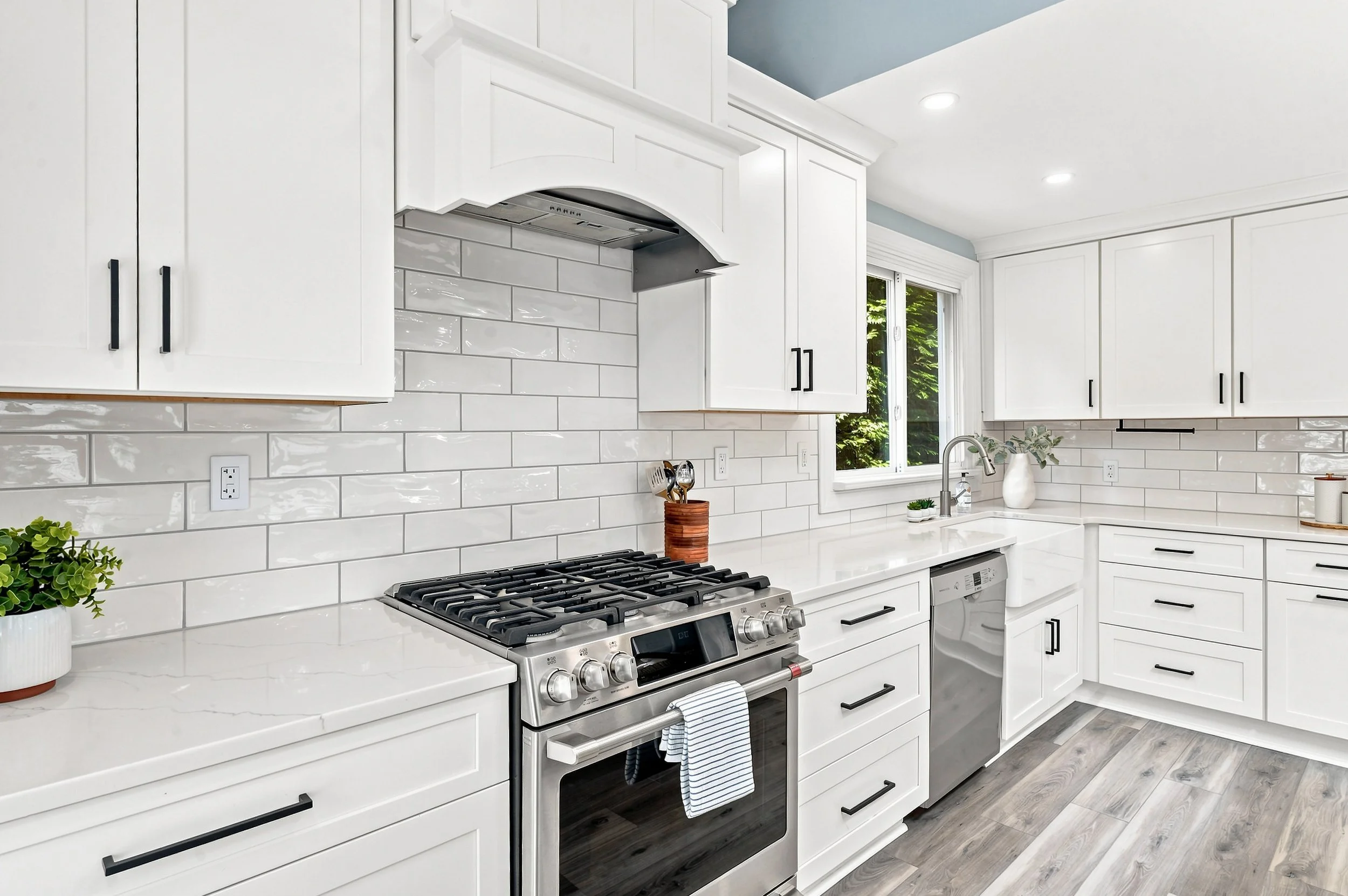 A modern kitchen with white cabinetry, a white countertop, a stainless steel gas stove, and a white tile backsplash. There is a window with green trees outside, and a potted plant on the counter.
