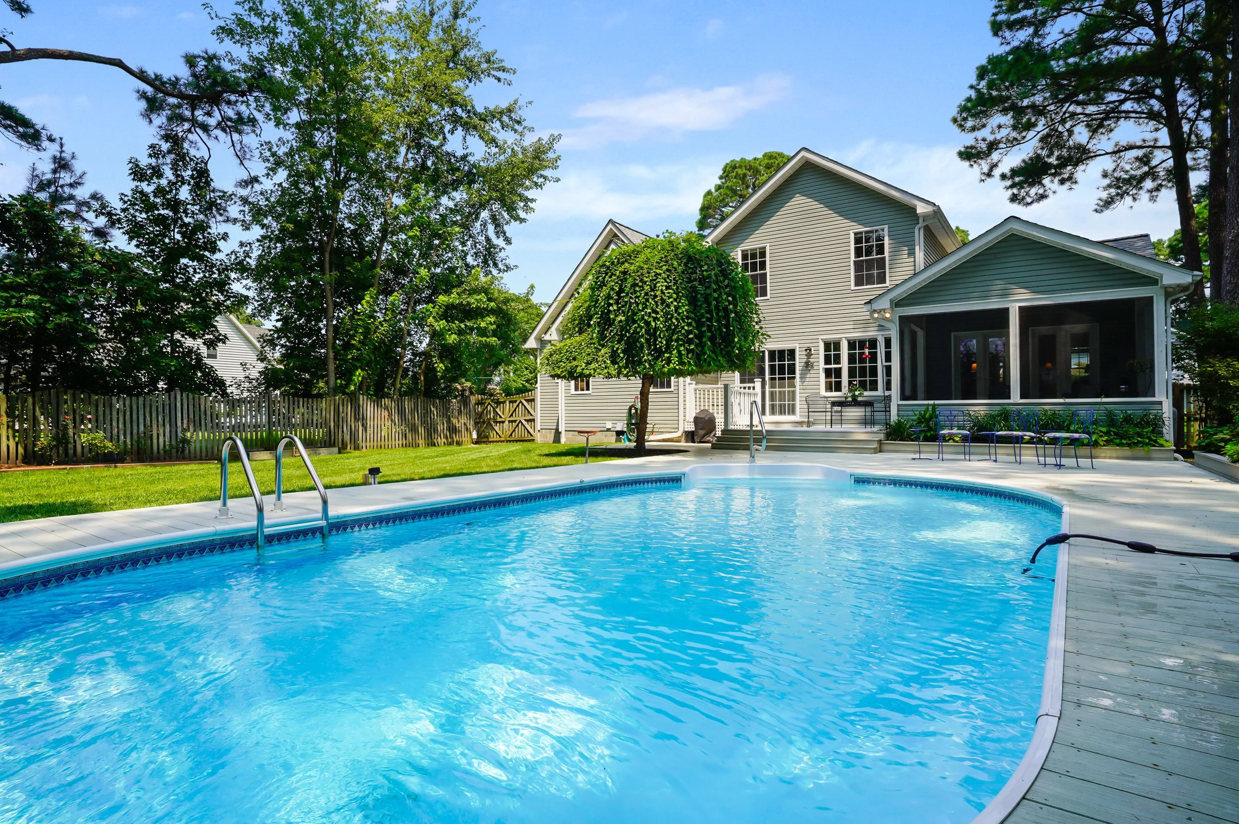 A backyard with a swimming pool, surrounded by a wooden fence and green trees, with a two-story house in the background and a bright, partly cloudy sky.