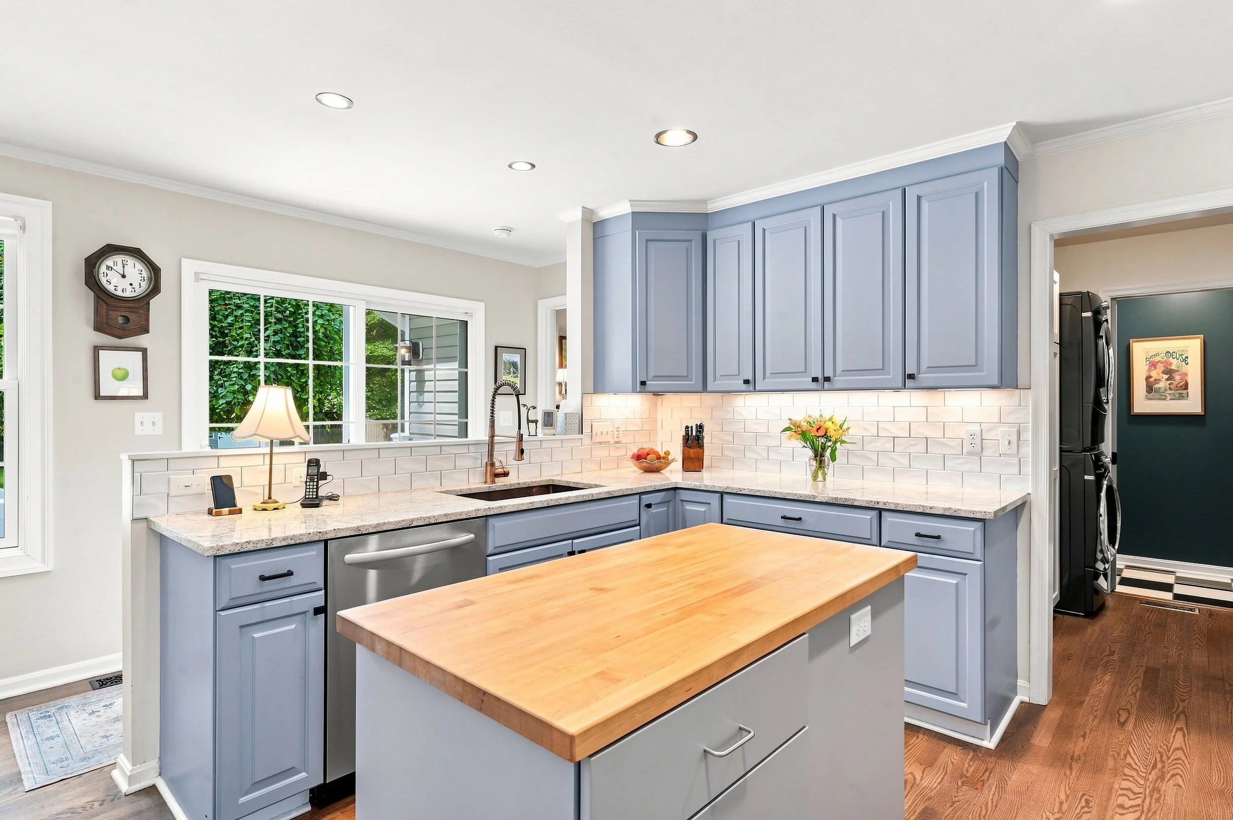 Kitchen with blue cabinets, white subway tile backsplash, wooden island, large window, and various small appliances and decorations.