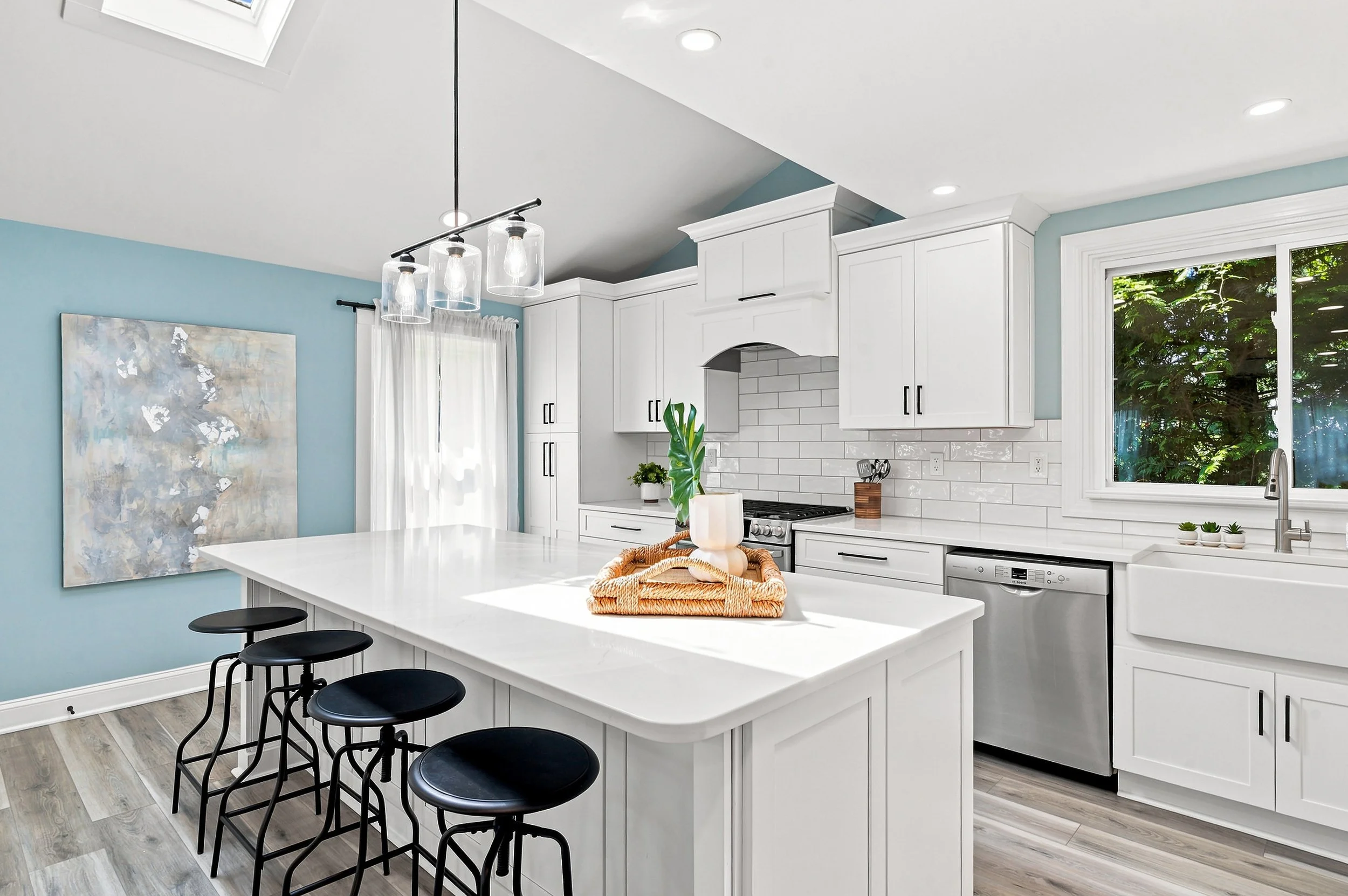 A modern kitchen with white cabinets, a white subway tile backsplash, and a kitchen island with black barstools. There is a window above the sink showing greenery outside. The kitchen has light gray wood flooring, a large abstract painting on a blue 