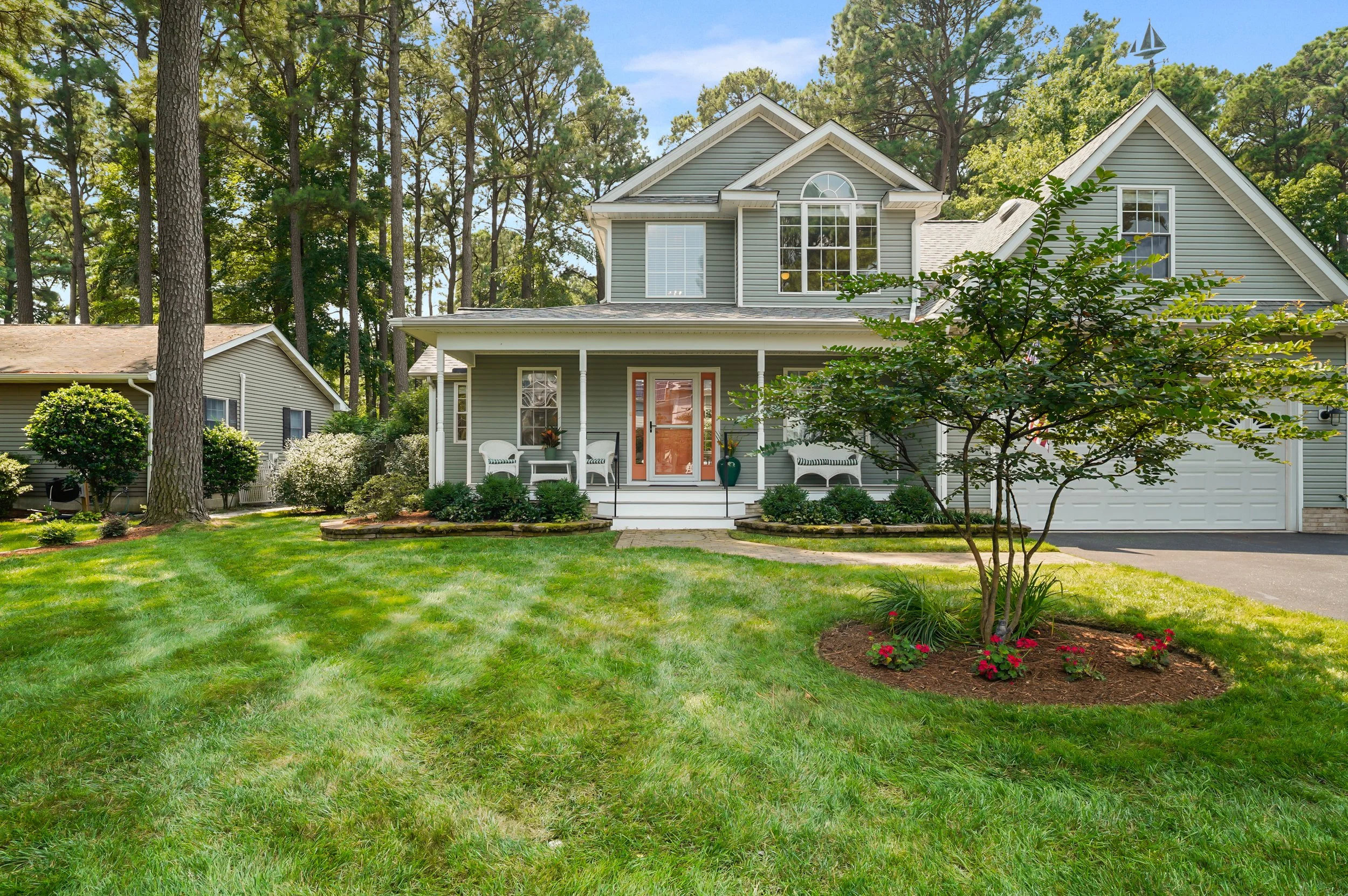 A two-story house with gray siding, white trim, and a front porch, surrounded by a green lawn, trees, and shrubs, under a bright blue sky.