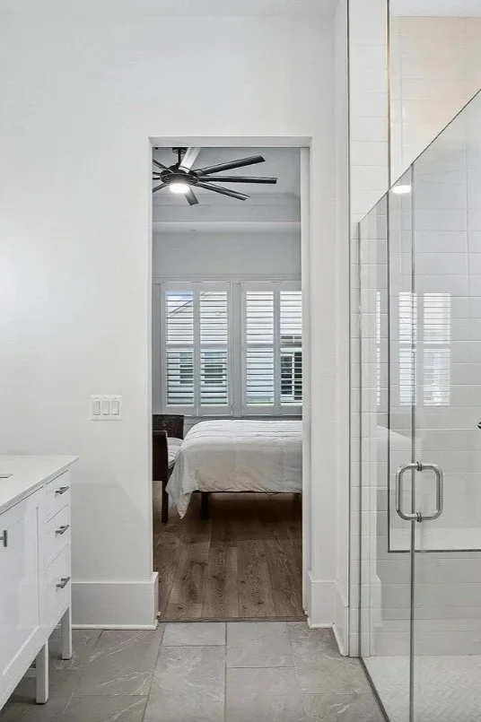 A doorway leading from a bathroom to a bedroom. The bedroom has a bed with white bedding, a ceiling fan, and windows with white shutters.