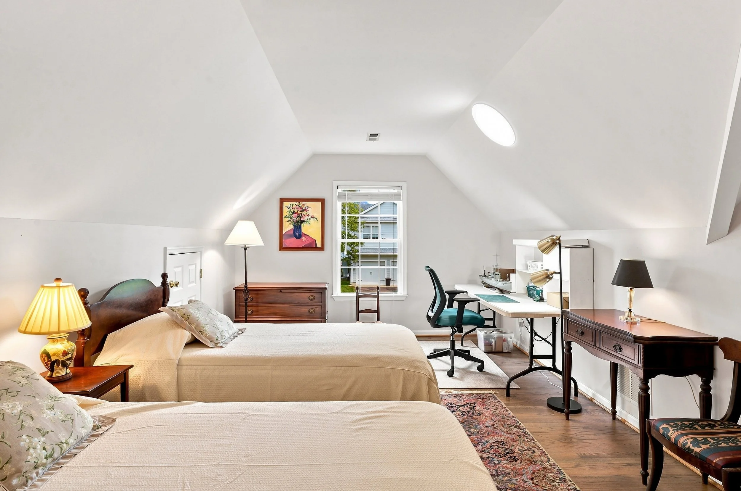A bedroom with two beds, nightstand lamps, a window, and a workspace with a chair, desk, and decorative items.