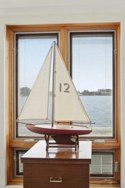 Model sailboat with the number 12 on its sail, displayed on a wooden table in front of a large window overlooking a body of water.