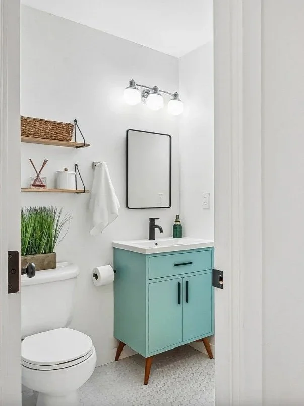 Small bathroom with white walls, a white toilet, a blue vanity with black handles, a small sink, a black faucet, a mirror, and a wall light fixture with three bulbs. There are two wooden shelves, a basket, decorative items, a towel, a soap dispenser, and a small potted plant.