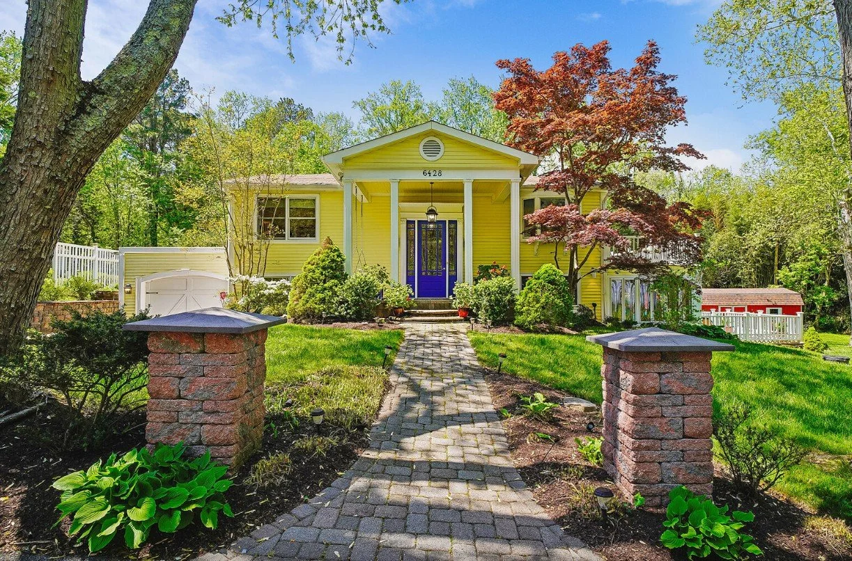 Yellow house with a blue front door, surrounded by green plants and trees, with a brick pathway leading to the front porch.