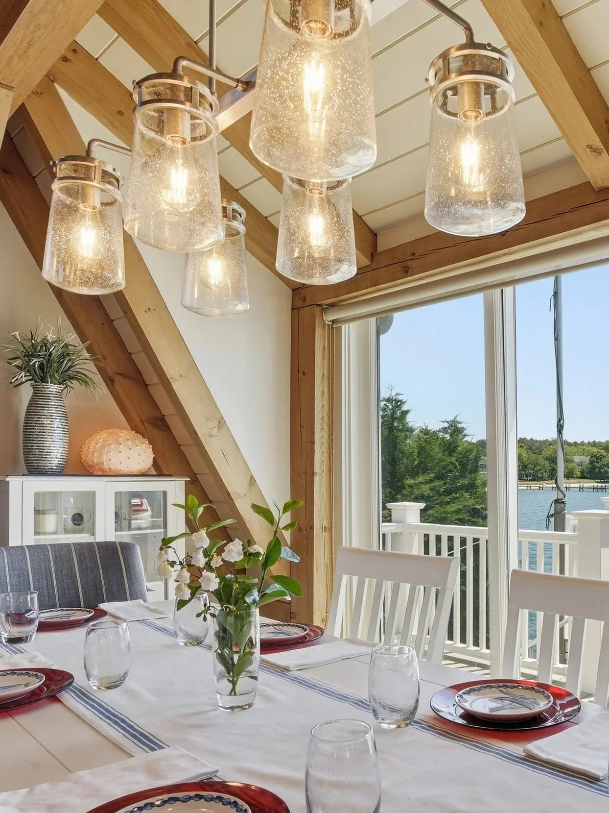 A dining room with a wooden-beamed ceiling and large window overlooking water and trees. A dining table is set with plates, glasses, and a vase with white flowers, with hanging glass and metal light fixtures above.