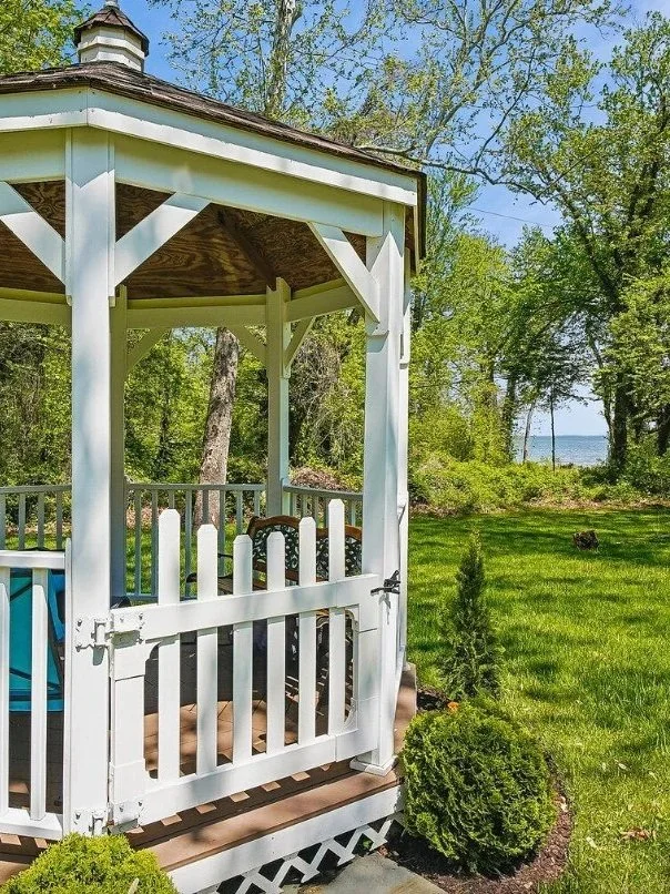A white wooden gazebo with a brown shingled roof, situated in a lush green garden near a body of water, with trees and shrubs surrounding it.