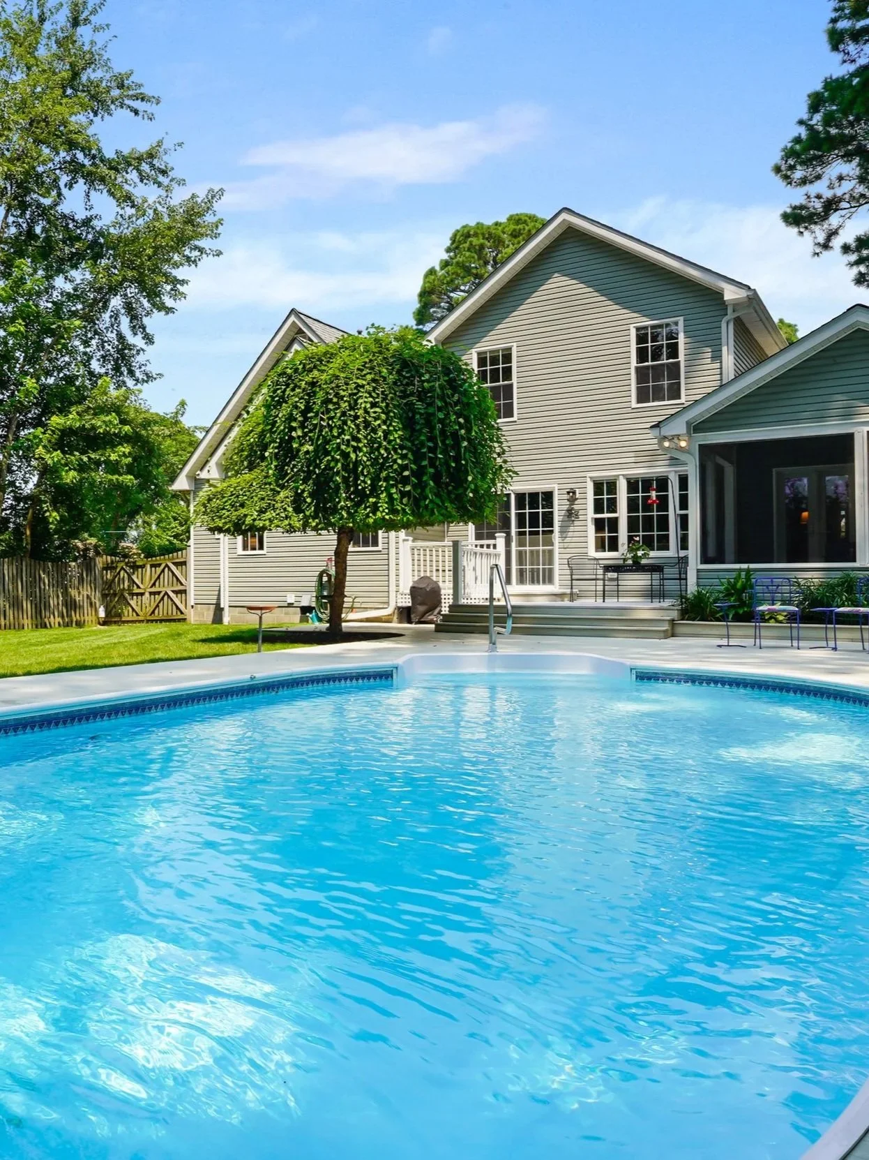 A backyard with a swimming pool, a tree, a house with a deck, and a well-maintained lawn and fencing.