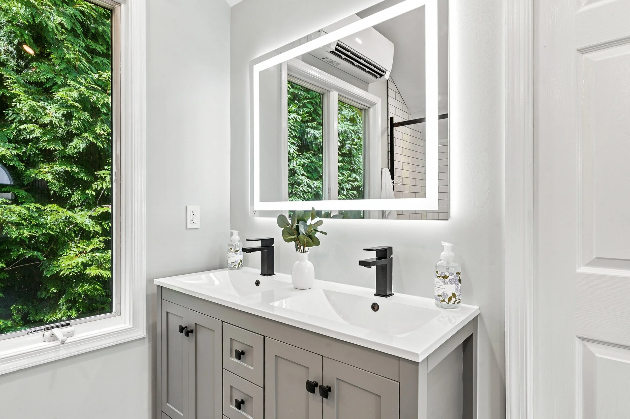 Bathroom with white walls, large window with greenery outside, white vanity with two black faucets, a potted plant, and a bottle of hand soap.