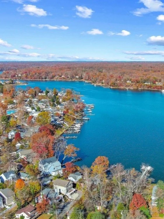 Aerial view of a lakeside community with houses and trees in fall colors, next to a large blue lake under a partly cloudy sky.