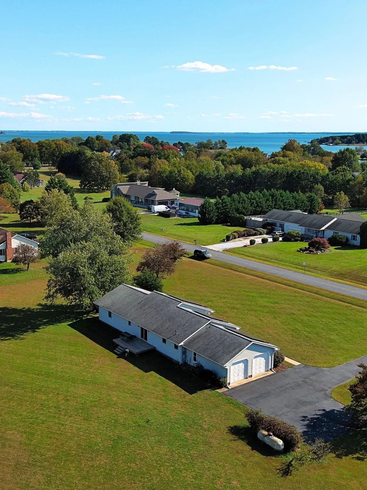Aerial view of a residential neighborhood near a body of water on a sunny day, with houses, trees, and green lawns.