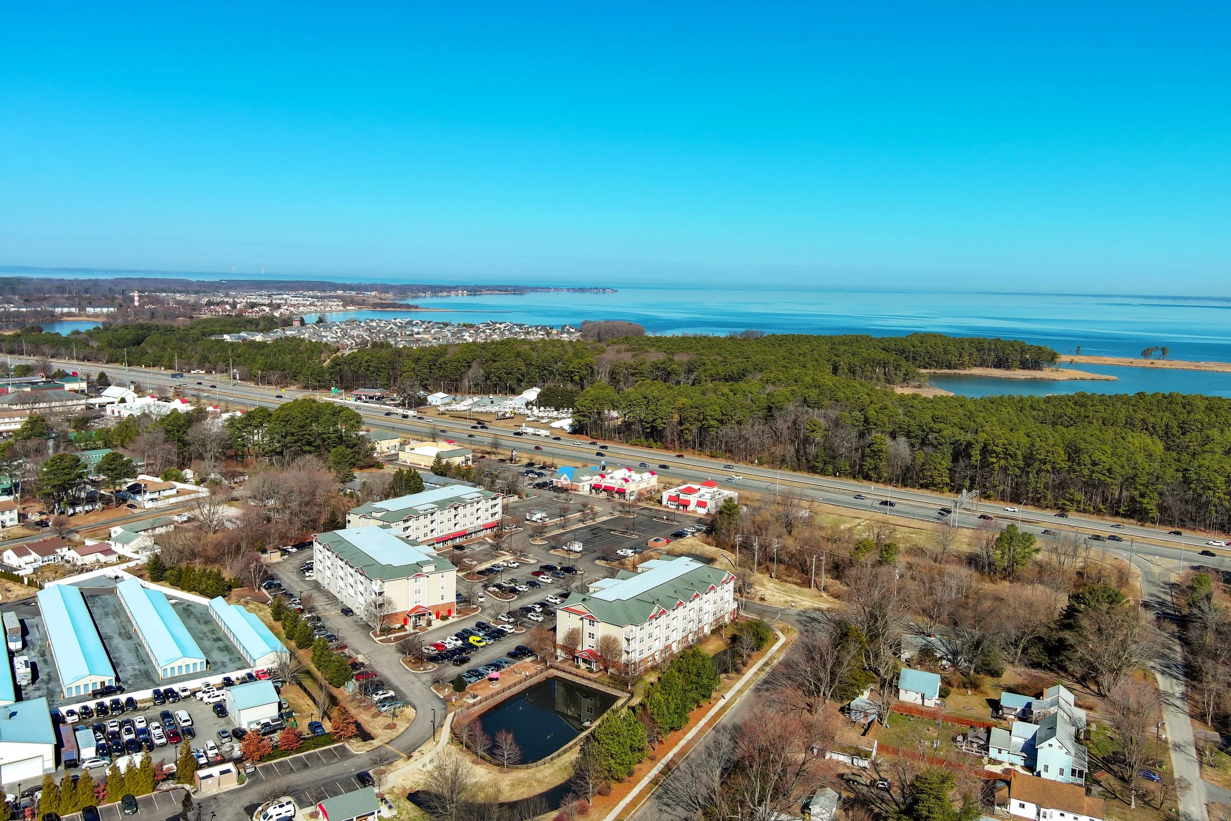 Aerial view of a coastal town with houses, trees, a parking lot, a highway, and a large body of water with a clear blue sky overhead.