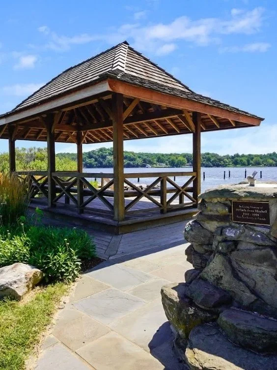 Wooden gazebo by a lake with a stone memorial plaque nearby.