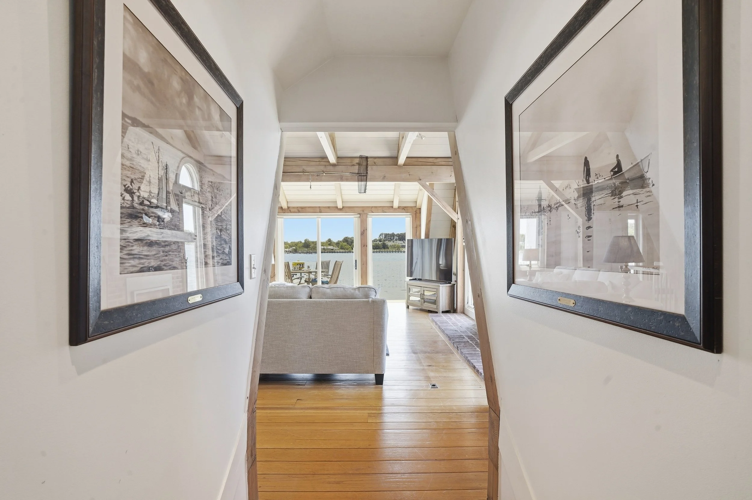 Inside view of a home with a wall hallway leading to an open living area with large windows showing an outdoor water view; framed artwork hangs on the hallway walls.