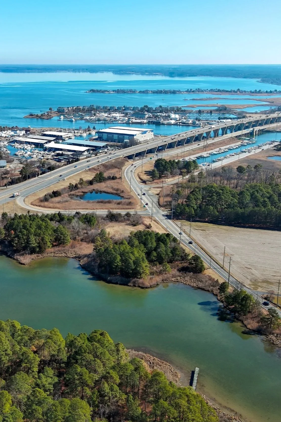Aerial view of a waterfront area with a bridge, marina, highway, and green lake surrounded by trees and land.