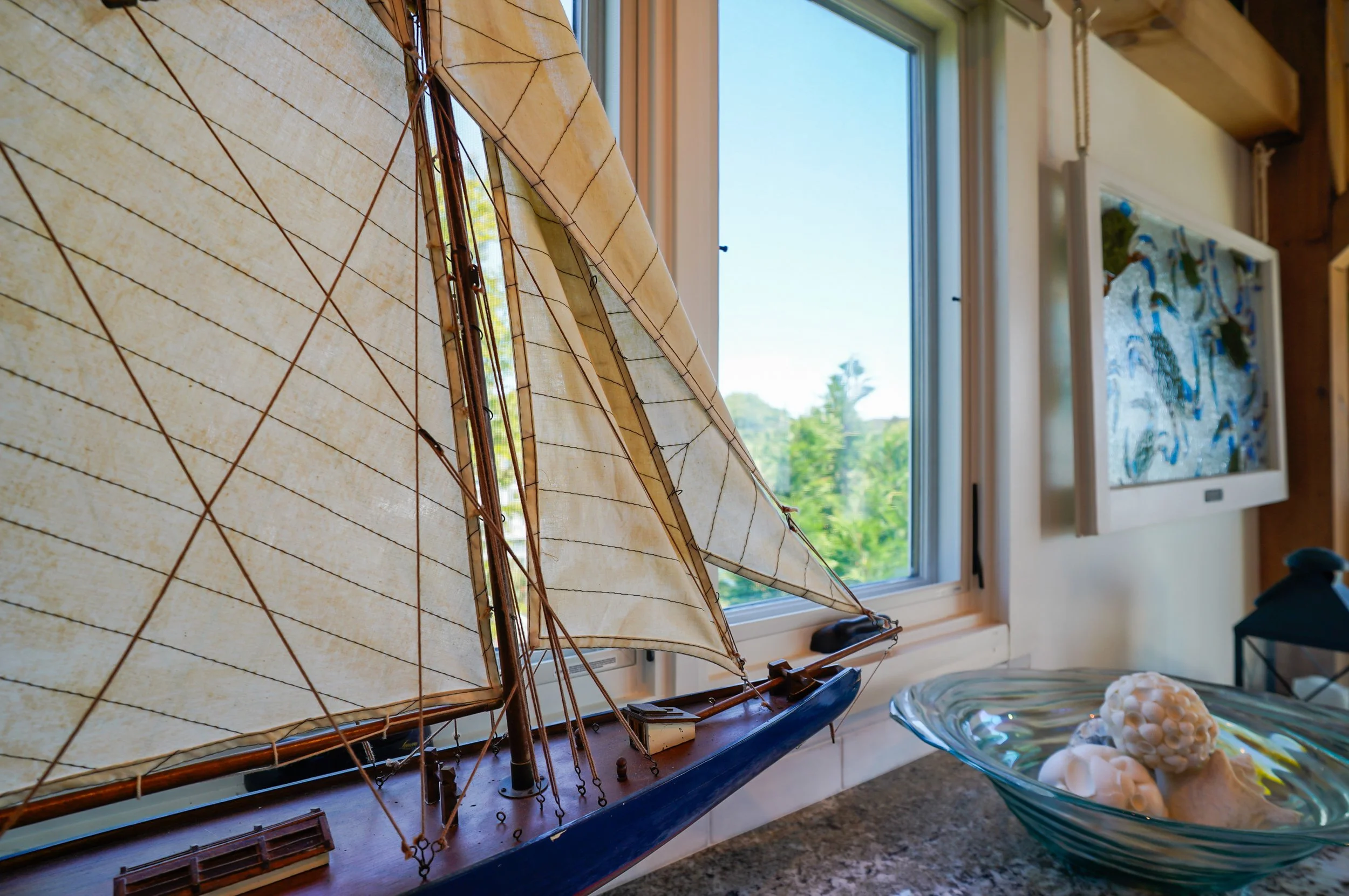 A model sailboat with a large cream-colored sail and wooden details is displayed on a windowsill inside a room. The window behind it shows a clear blue sky and green trees outside. To the right, there is a decorative artwork with blue and green patte