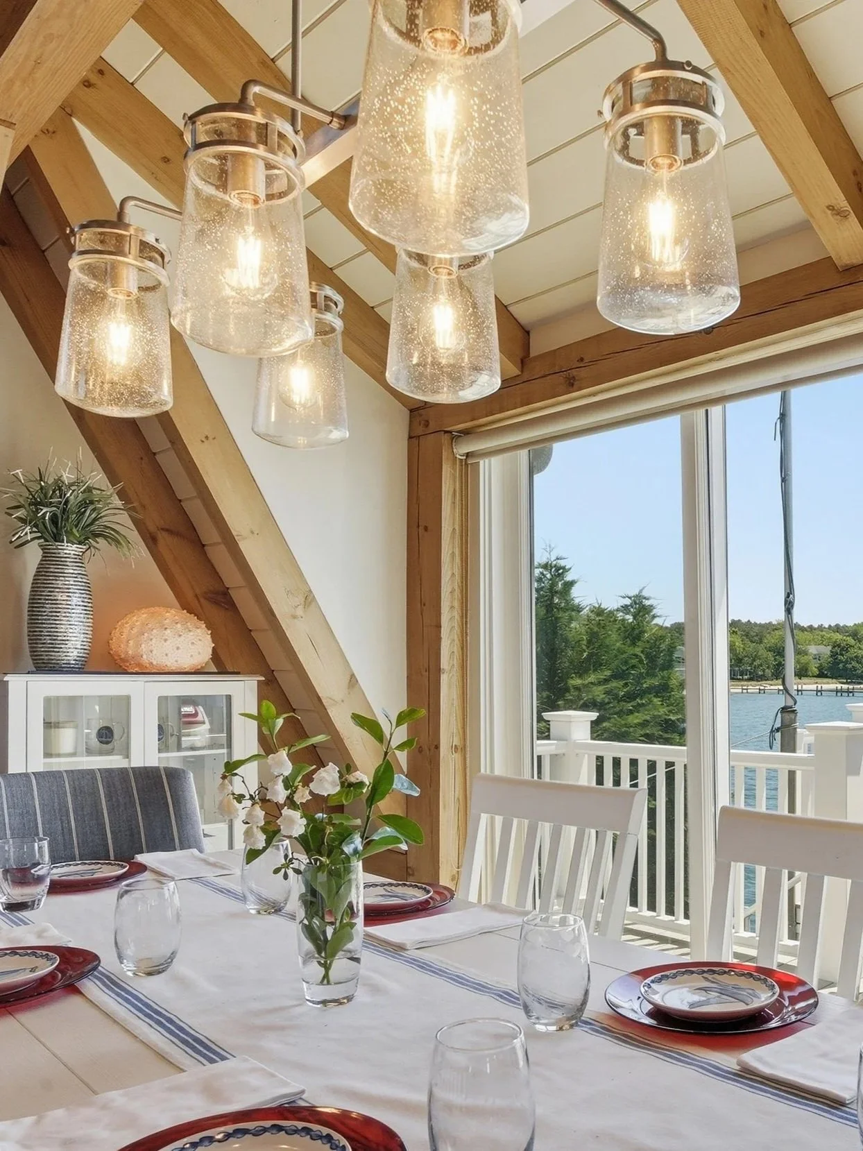 A dining room with a table set with plates, glasses, and a vase of white flowers. A wooden beam ceiling with hanging glass pendant lights, large window overlooking water, and outdoor deck railing.