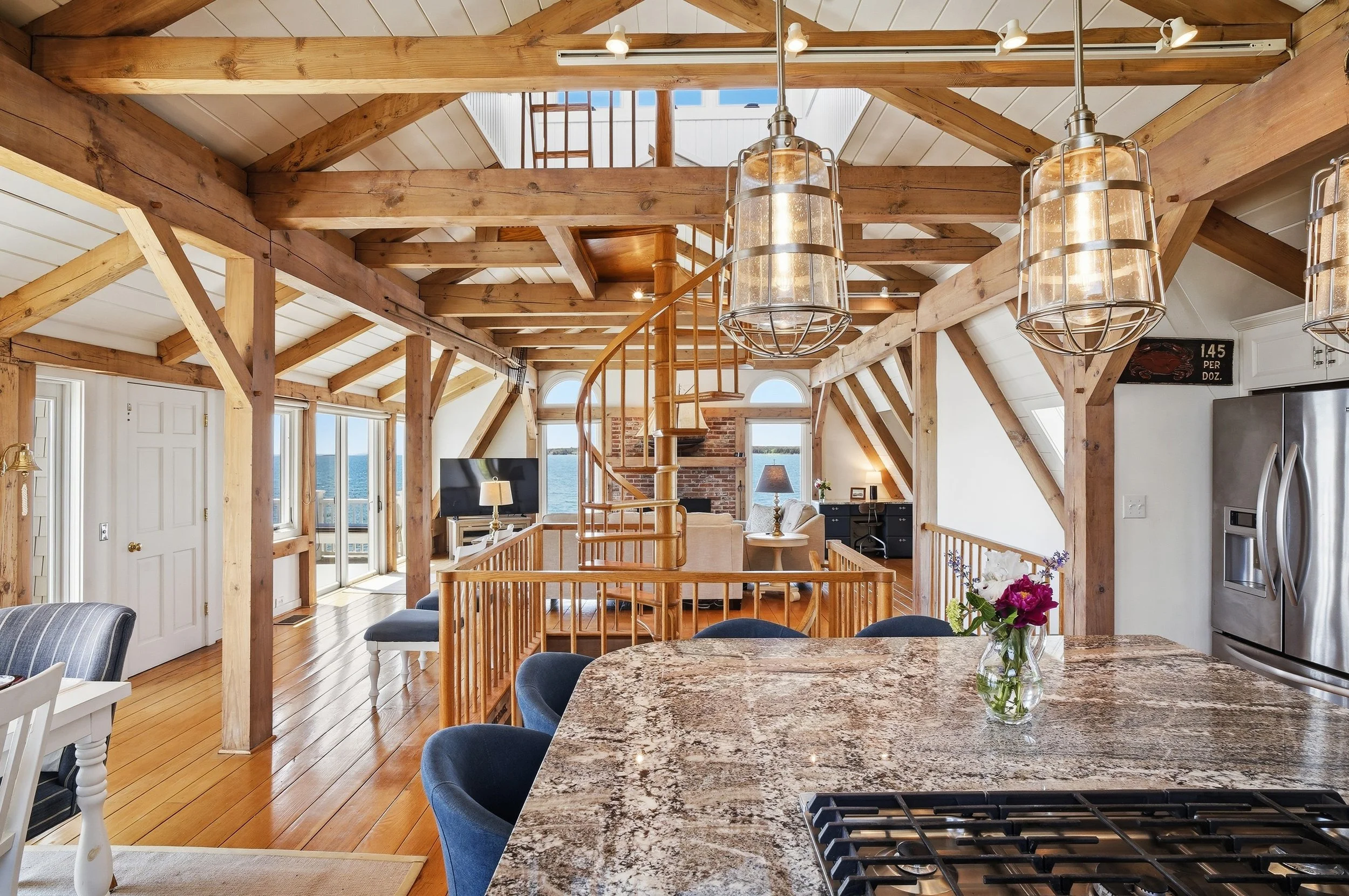 Interior view of a bright, spacious open-concept living area with wooden beams, a staircase, and large windows showing water and sky outside. There are hanging pendant lights, a kitchen with granite countertops, a dining table with a vase of flowers,