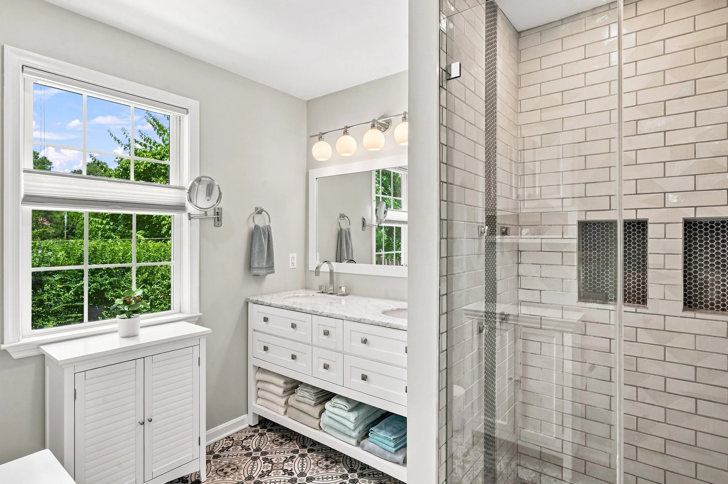 A bright bathroom with a large window showing green trees outside. There is a white vanity with drawers and shelves holding folded towels, a marble countertop, and a mirror above it. Thermometers and towel racks are mounted on the wall, and a window 