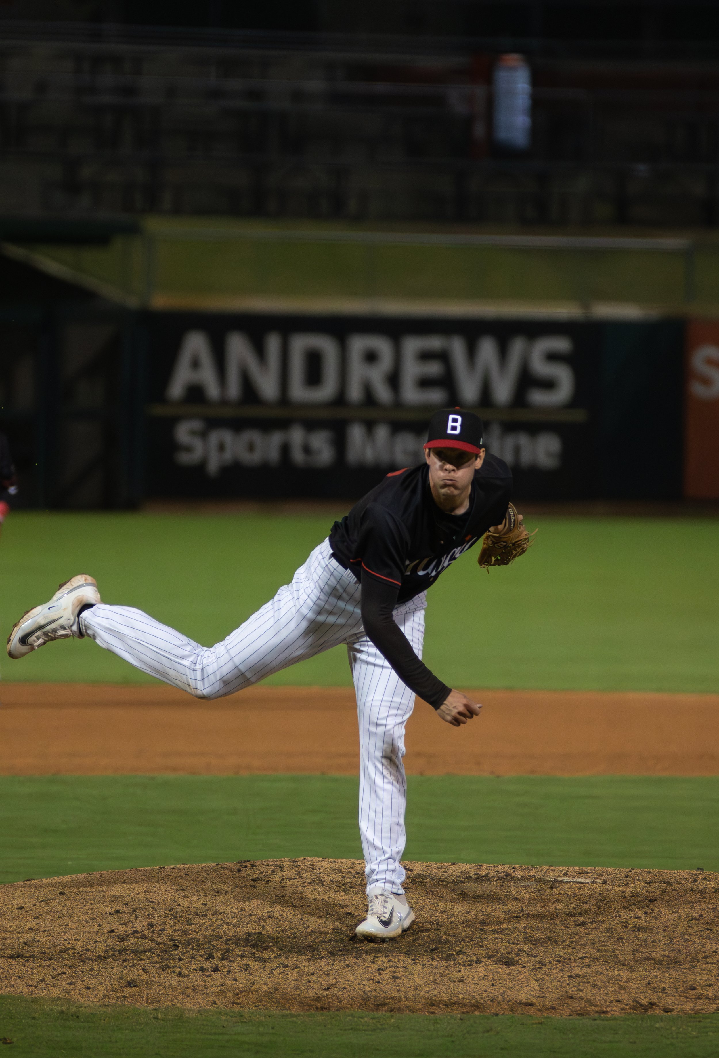 A professional baseball pitcher is in the middle of his pitch, on the mound at a baseball stadium, with an audience empty stadium seats in the background.