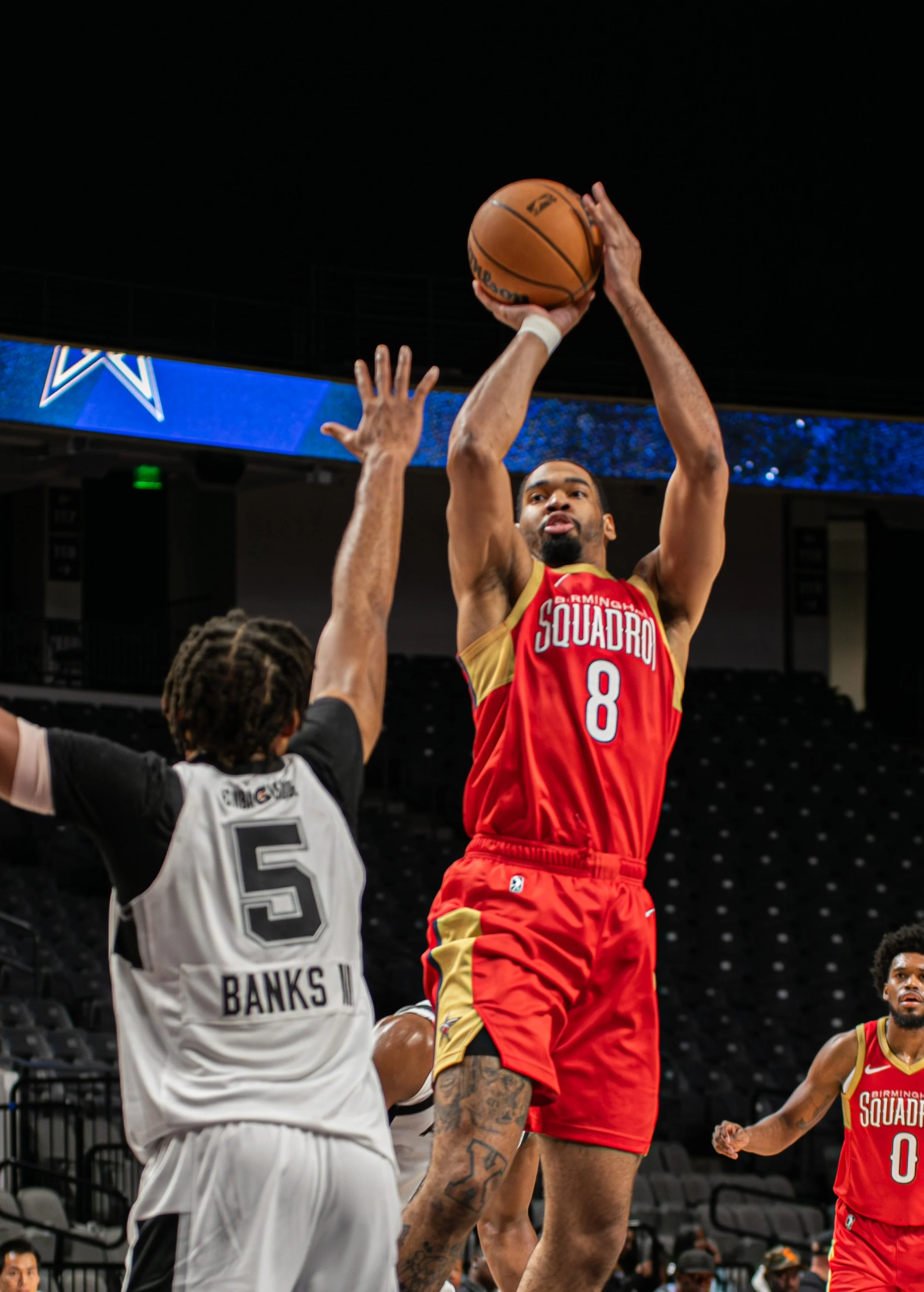 A basketball player in a red and yellow uniform jumping to shoot the ball while an opponent in a black and white uniform attempts to block the shot on an indoor court.