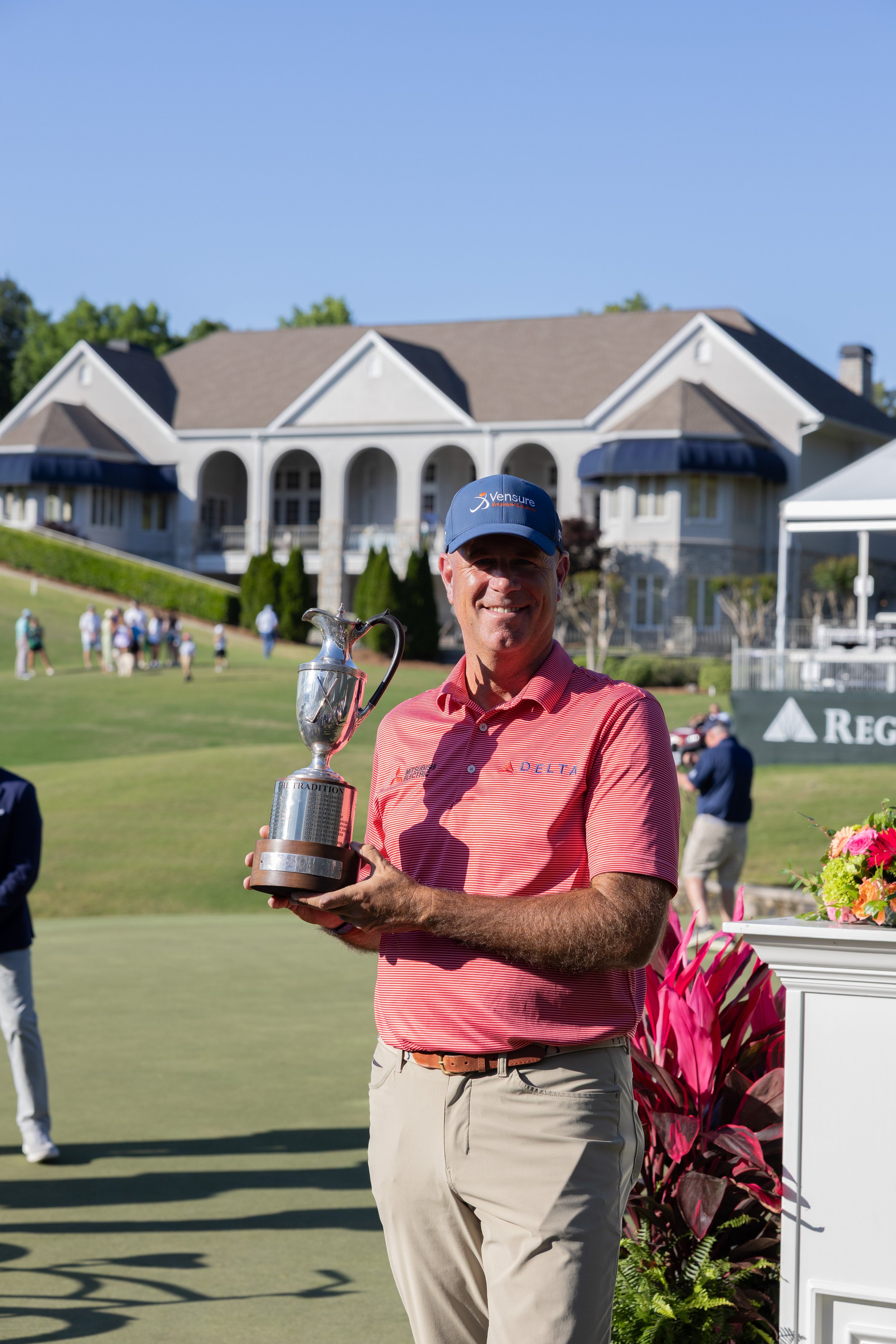 Man smiling and holding a silver trophy on a golf course with a large house in the background and people in the distance.