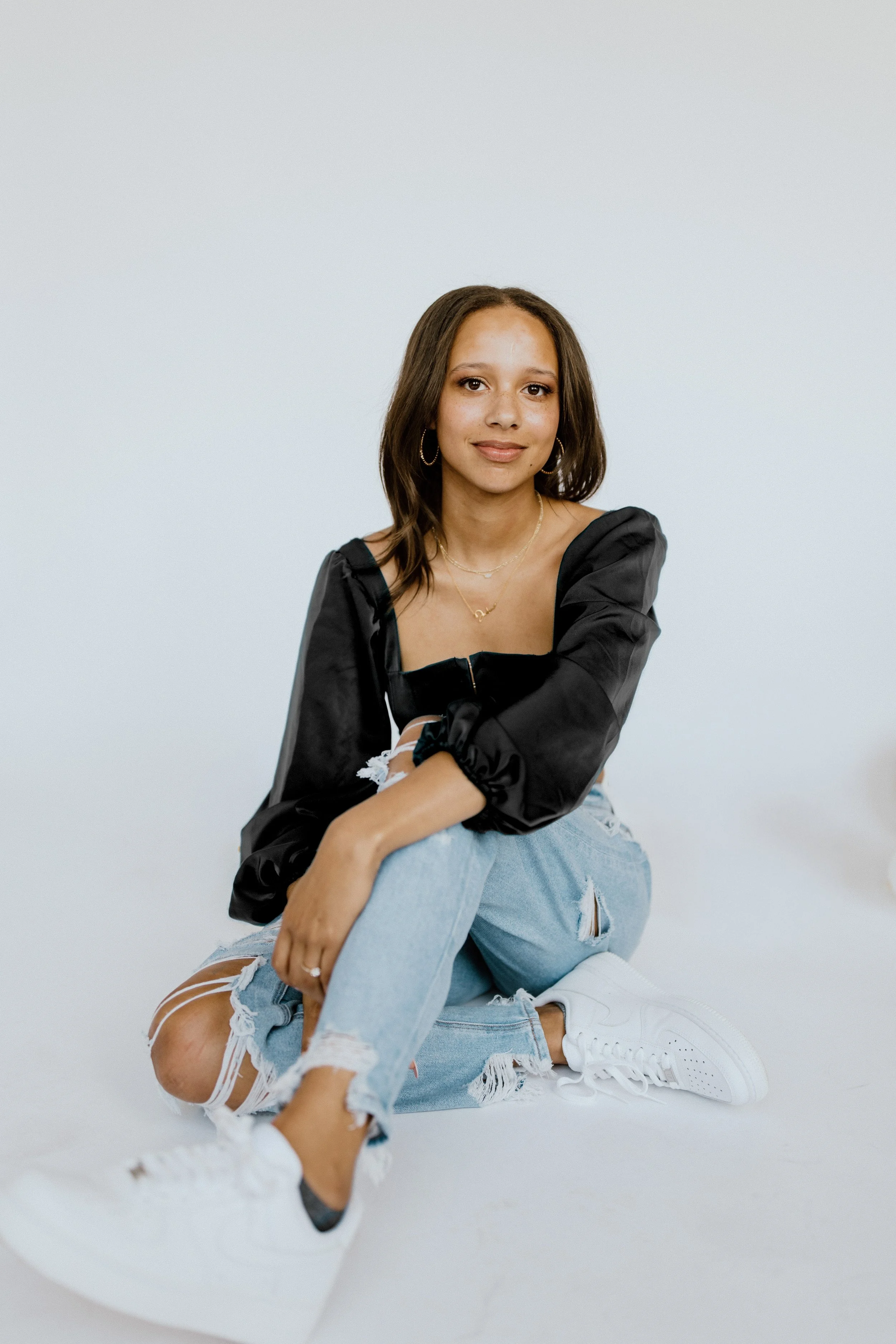 Young woman sitting on the floor in front of a plain white wall, wearing a black top, ripped light blue jeans, white sneakers, and hoop earrings, smiling softly at the camera.
