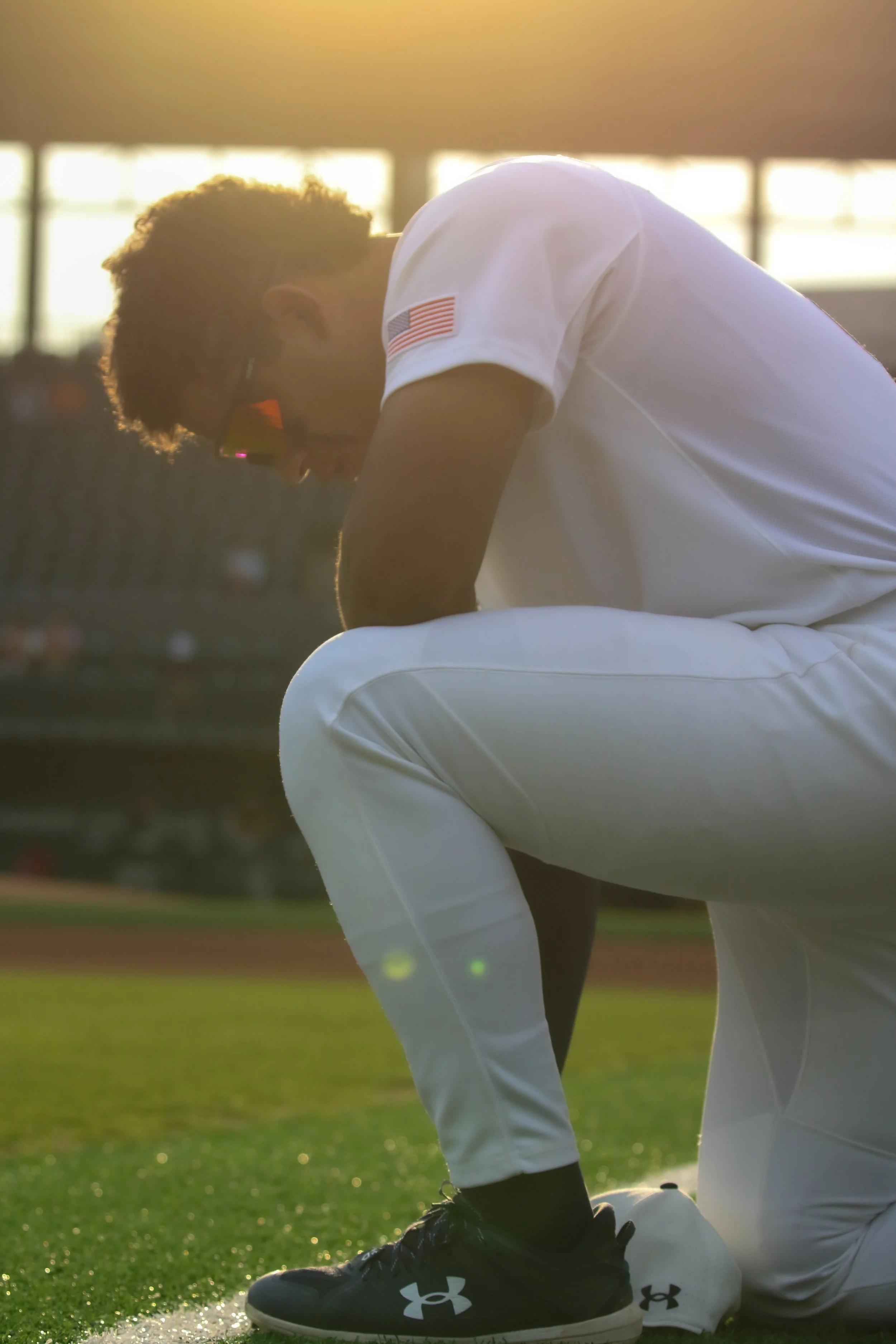 A young athlete in a white sports uniform with an American flag patch on the sleeve, kneeling on a green field at sunset, with the sun backlighting the scene.