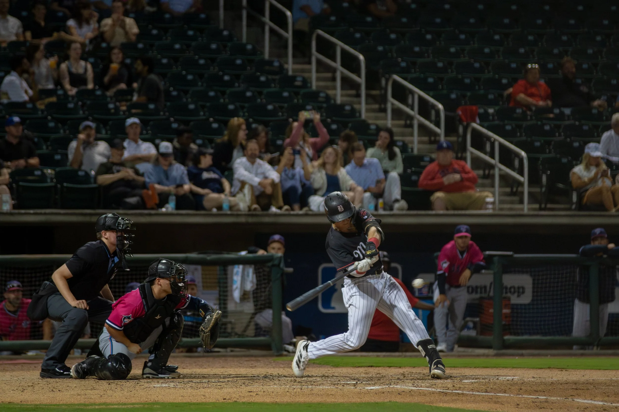 A baseball game in progress with a player sliding into home plate as the catcher and umpire watch. Spectators are seated in the background, some taking photos.