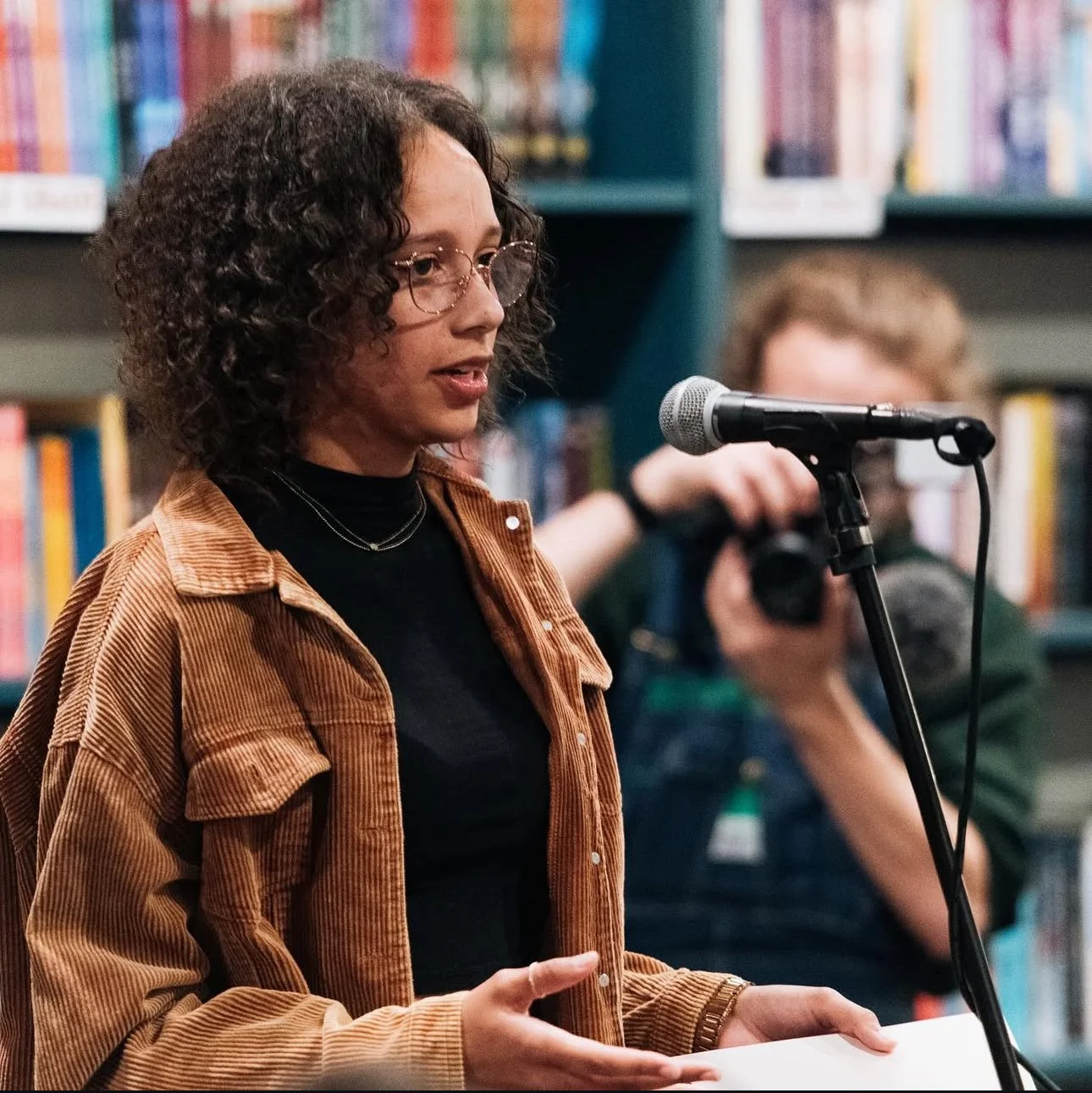 A woman with curly hair and glasses speaks into a microphone at a library. She is wearing a brown corduroy jacket over a black top. In the background, a person with a camera records her, with bookshelves behind them.