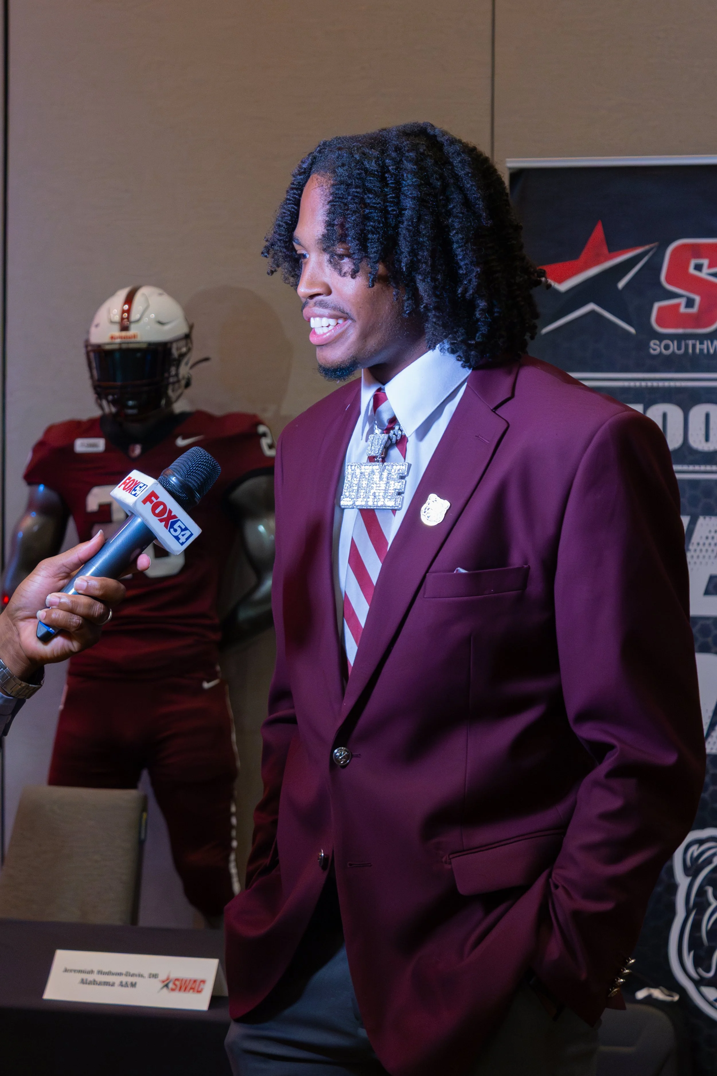 A man in a maroon suit and tie being interviewed by a news reporter at a press event, with a football player in a crimson jersey and helmet in the background.