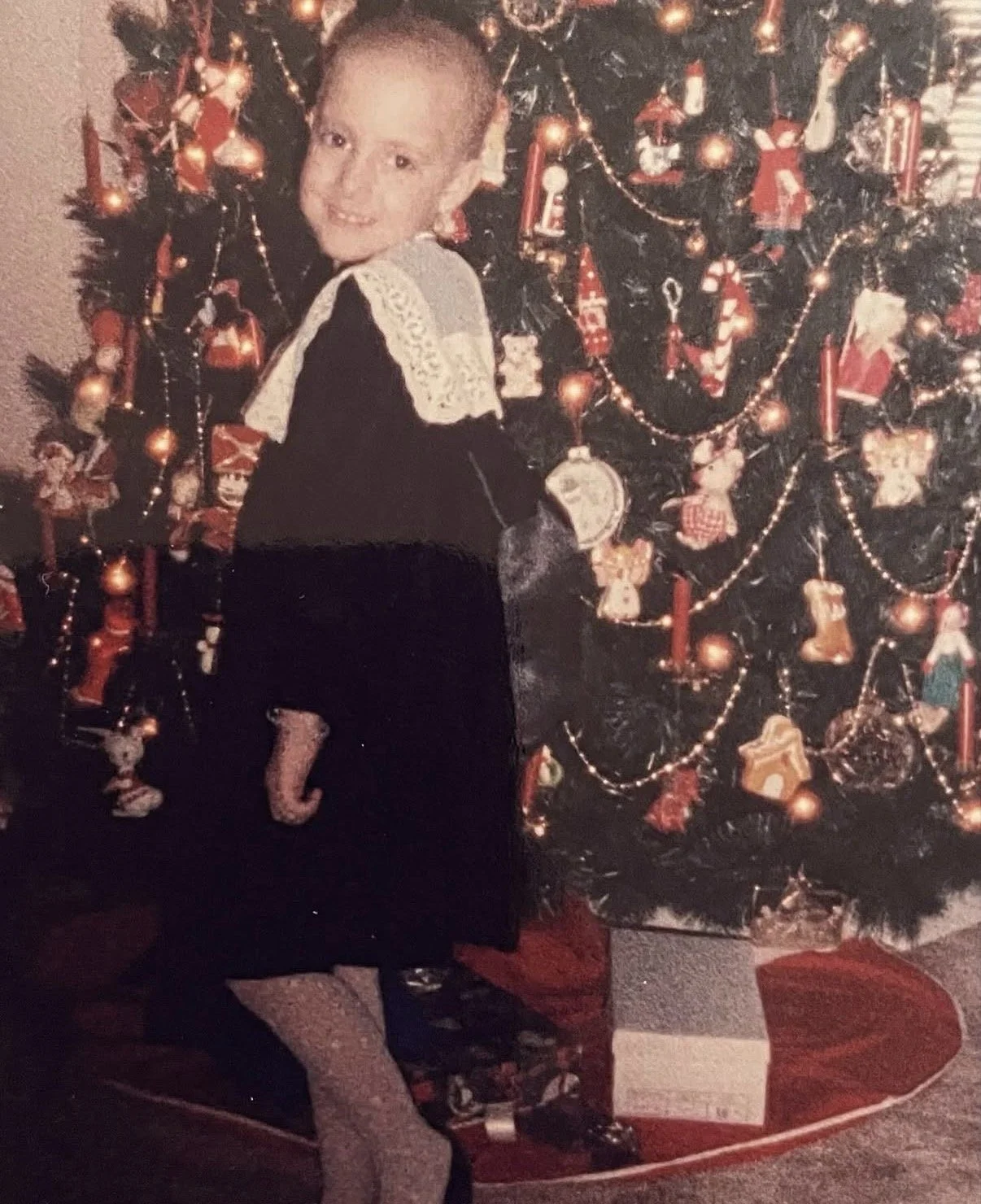 A young boy with a shaved head smiling and standing in front of a decorated Christmas tree, wearing a black dress with a white lace collar, colorful tights, and black shoes with bow embellishments.