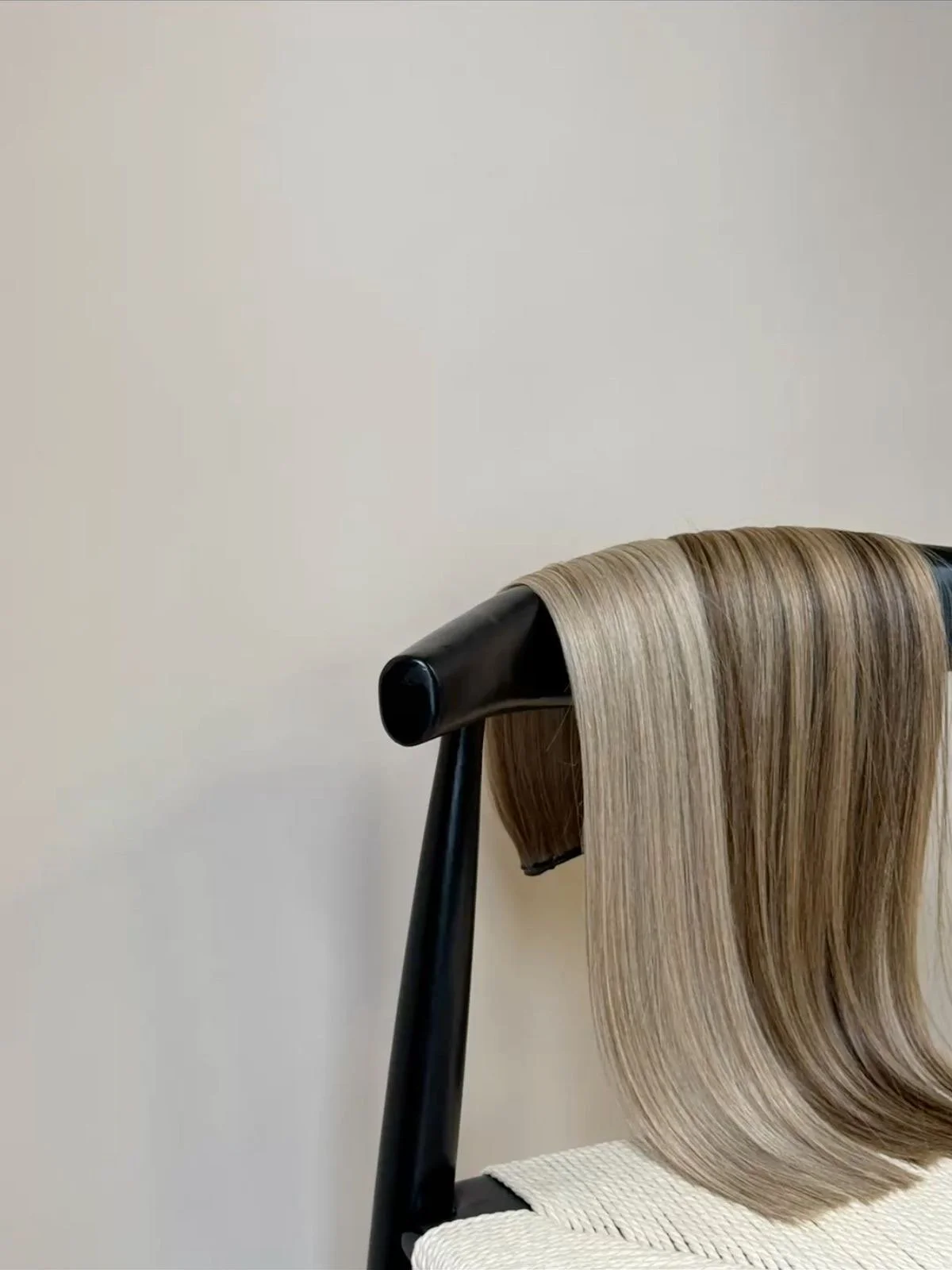 A black chair with a beige cushion, holding long, straight, blonde hair draped over the backrest, against a plain off-white wall.