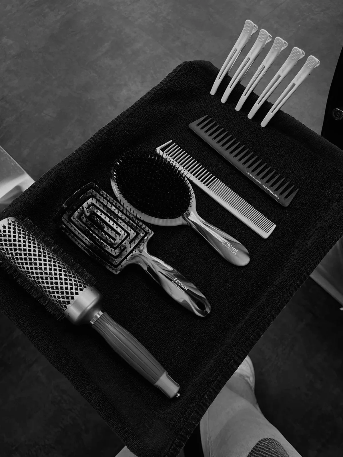 Assorted hair styling tools including a round brush, hair grater, hair comb, wide-tooth comb, and hair clips arranged on a black towel.