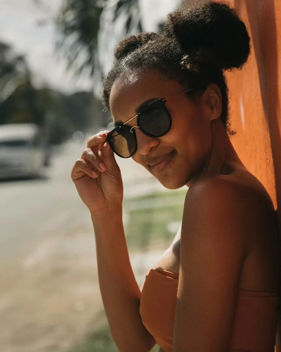 A woman with natural hair in two buns wearing sunglasses, smiling, and adjusting her glasses outdoors.