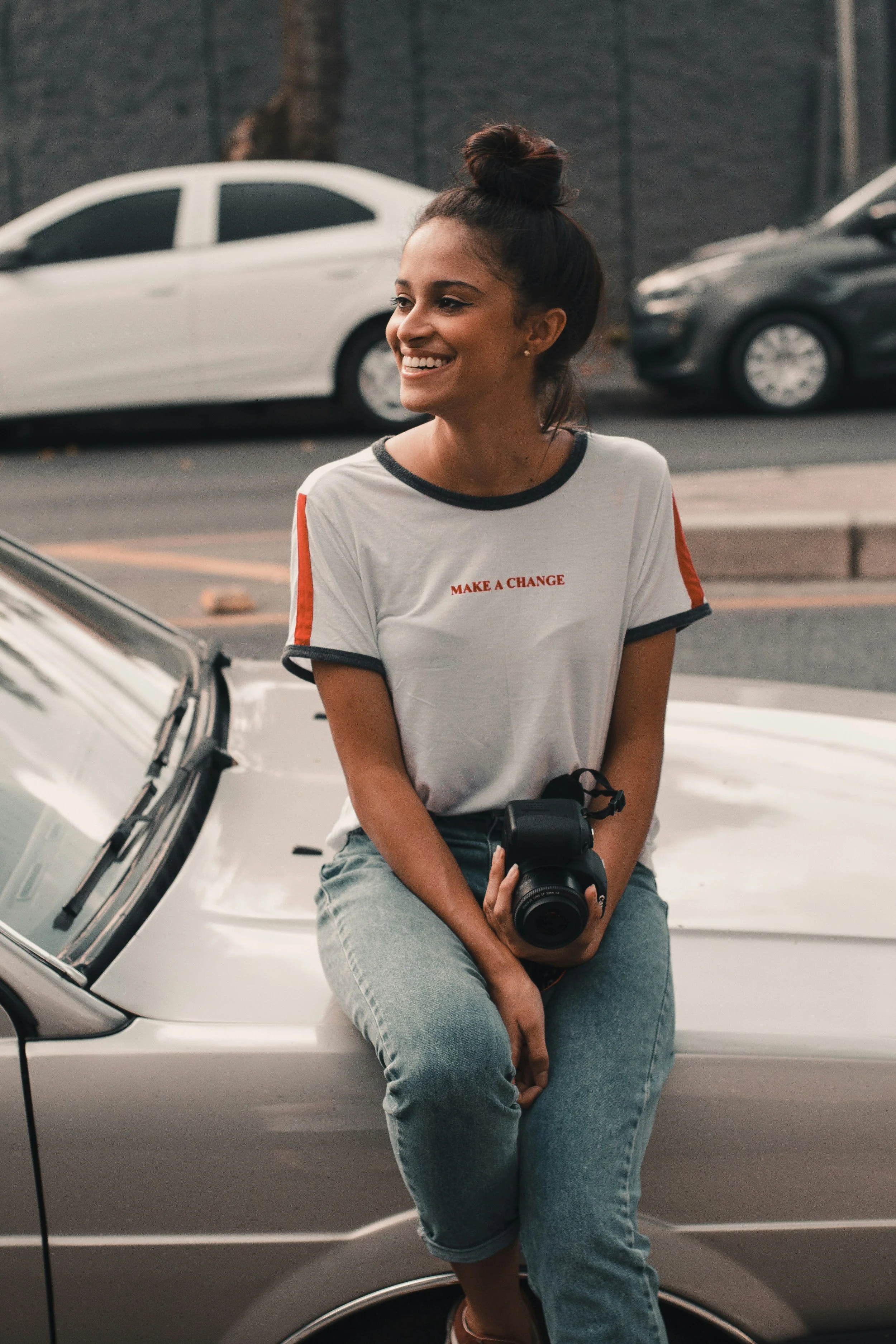 A young woman with dark hair in a bun, wearing a white T-shirt with red and black accents that says 'Make a Change,' is sitting on the hood of a silver car, holding a camera, smiling, in an outdoor parking lot with other cars and a black brick wall in the background.