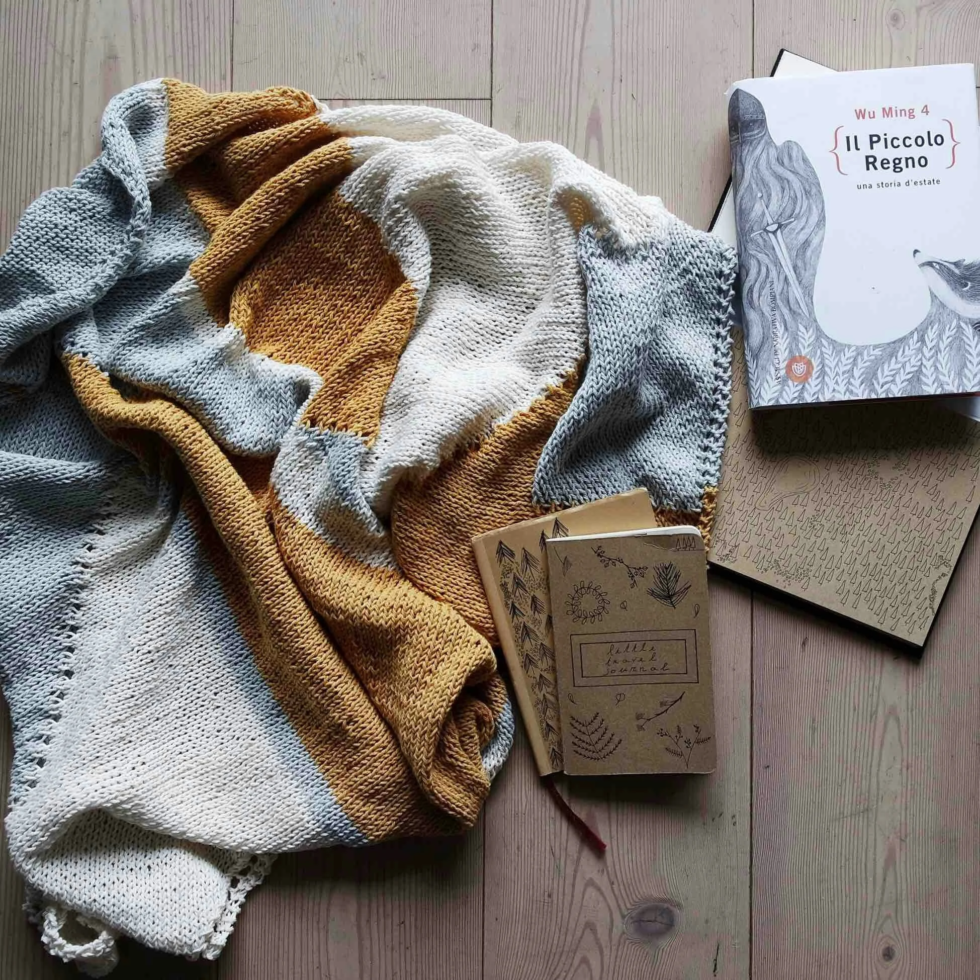 Colorful knitted blankets, a book titled "Il Piccolo Regno" by Wu Ming 4, and a few notebooks with plant illustrations are arranged on a wooden surface.