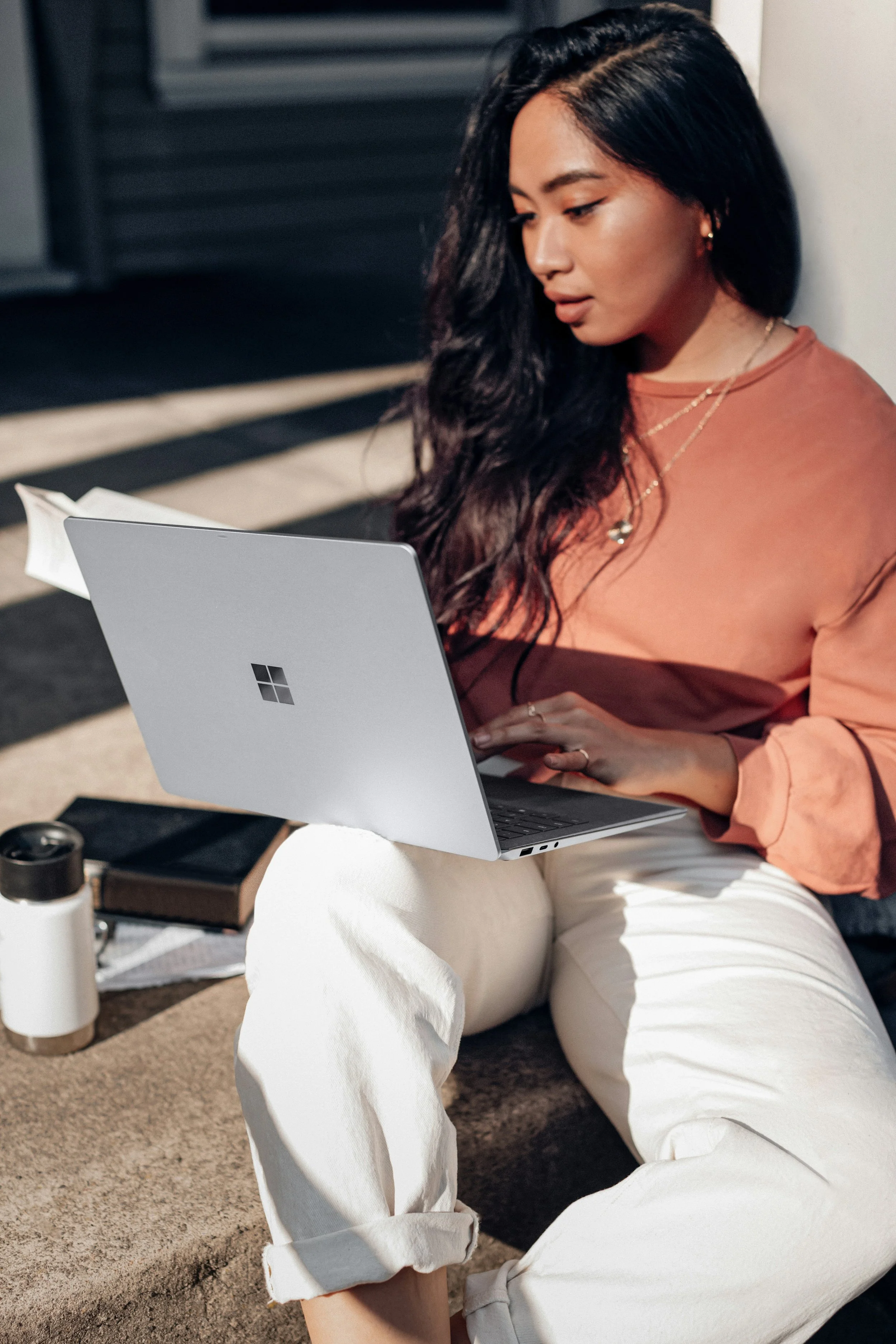 A woman sitting on the ground cross-legged, using a silver laptop, outdoors near a brick building, holding the laptop with her left hand, wearing a beige shirt and white pants, with a notebook and water bottle nearby.