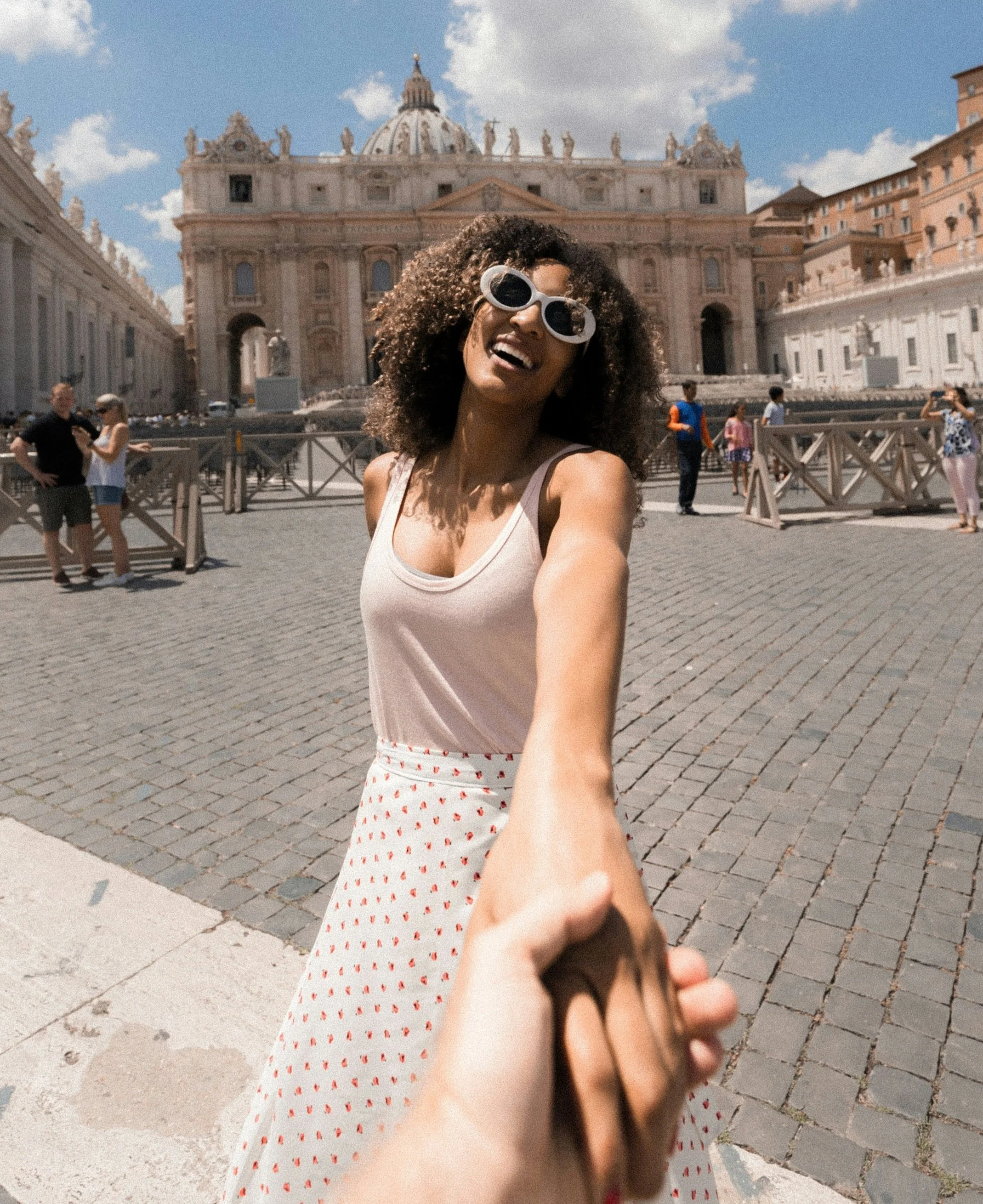 A woman with curly hair and large white sunglasses smiling for a selfie in front of a historic building with a dome, possibly St. Peter's Basilica in Vatican City, during daytime with partly cloudy sky.