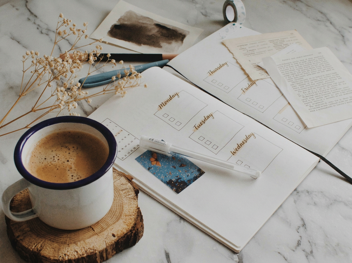 Open planner with weekly schedule, a cup of coffee on a wooden coaster, dried flowers, a pen, washi tape, and printed papers on a marble surface.