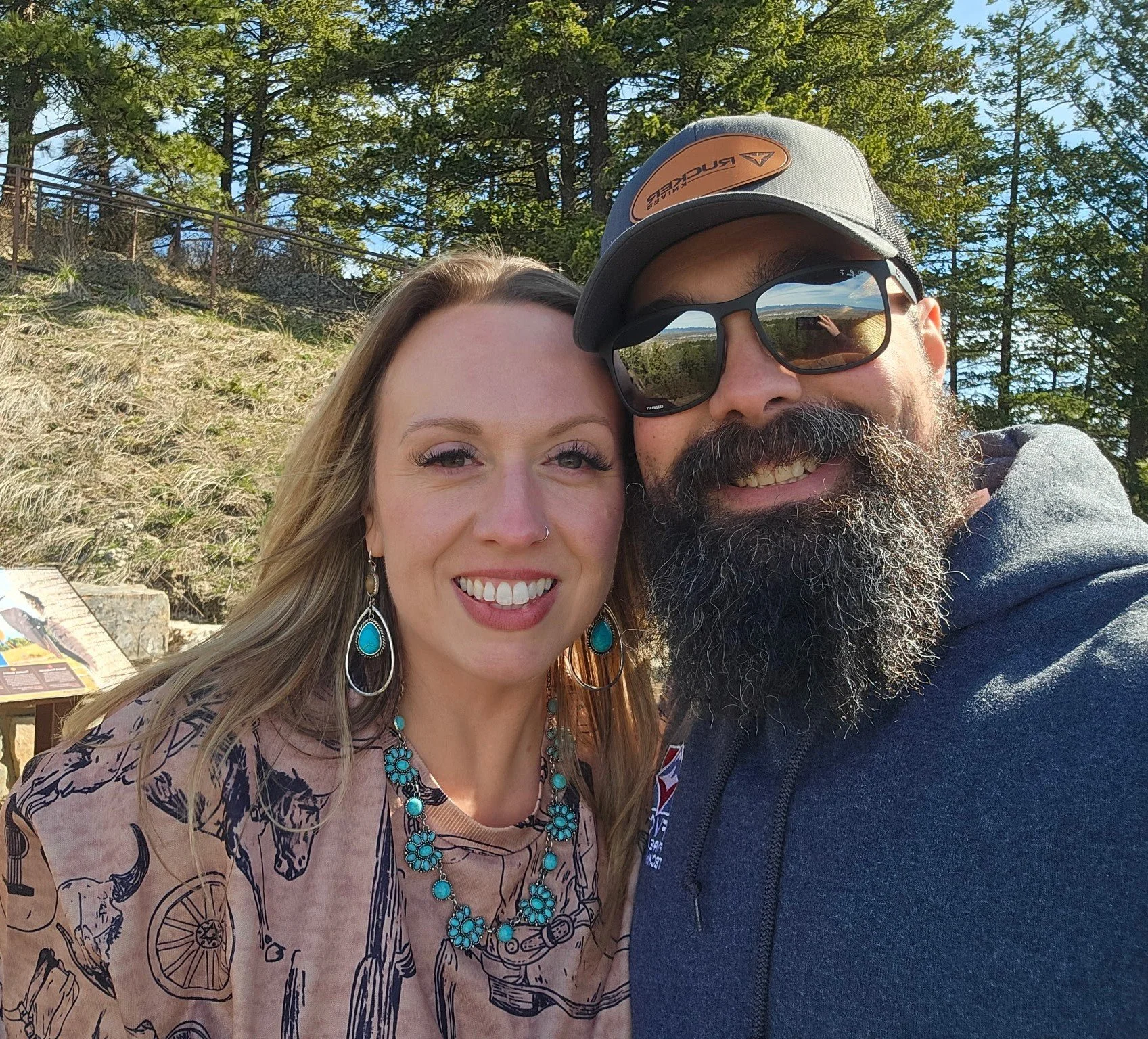 A smiling woman with long blonde hair, turquoise jewelry, and a nose ring stands next to a smiling man with a beard, sunglasses, and a gray baseball cap. They are outdoors with trees and a hill in the background.