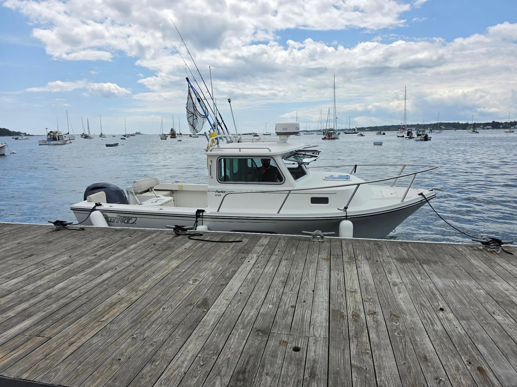 A white boat docked at a wooden pier on a body of water with several other boats in the background under partly cloudy skies.